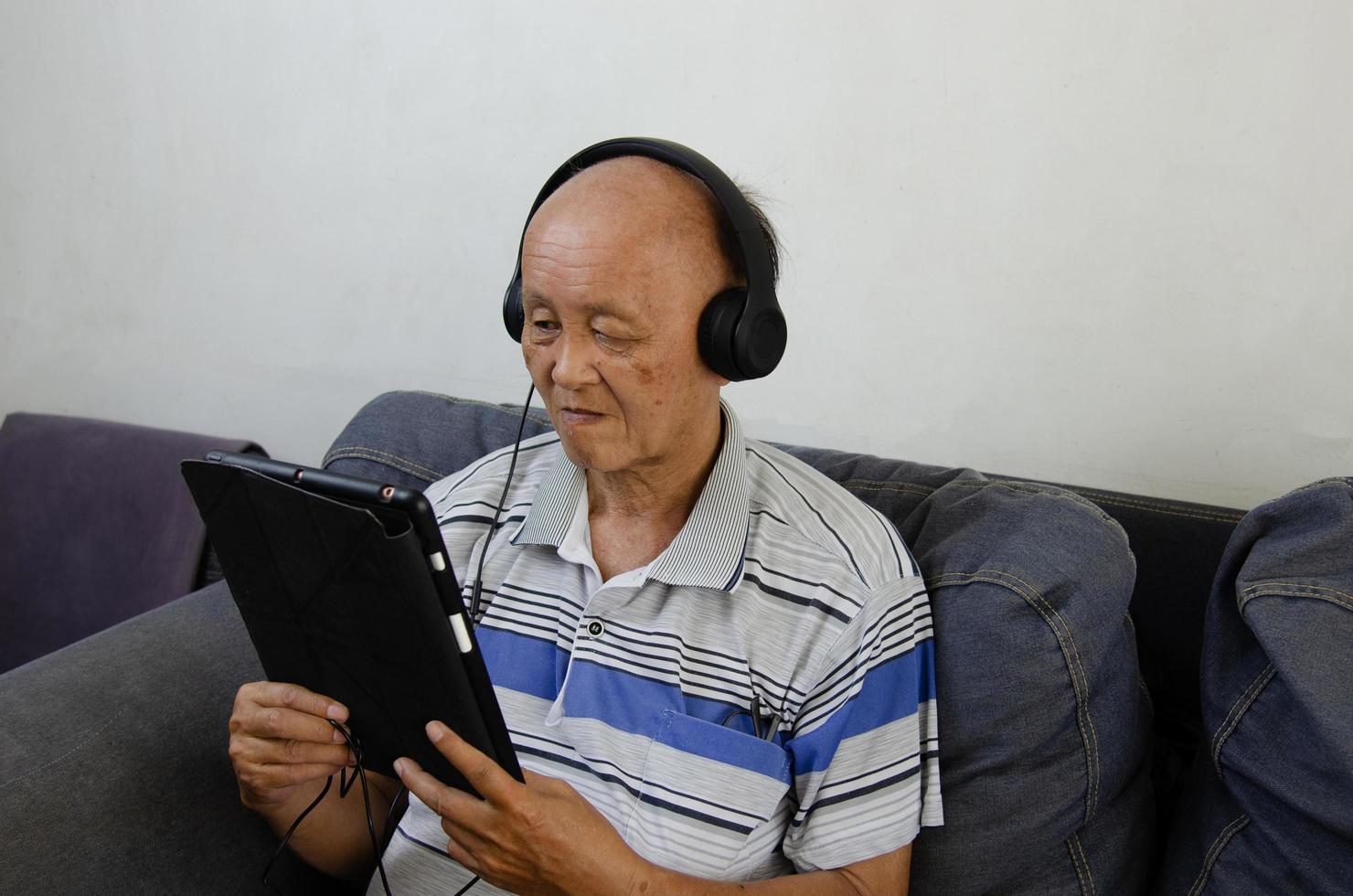 anciano sosteniendo una tableta en el sofá escuchando música con auriculares y medios sociales. foto