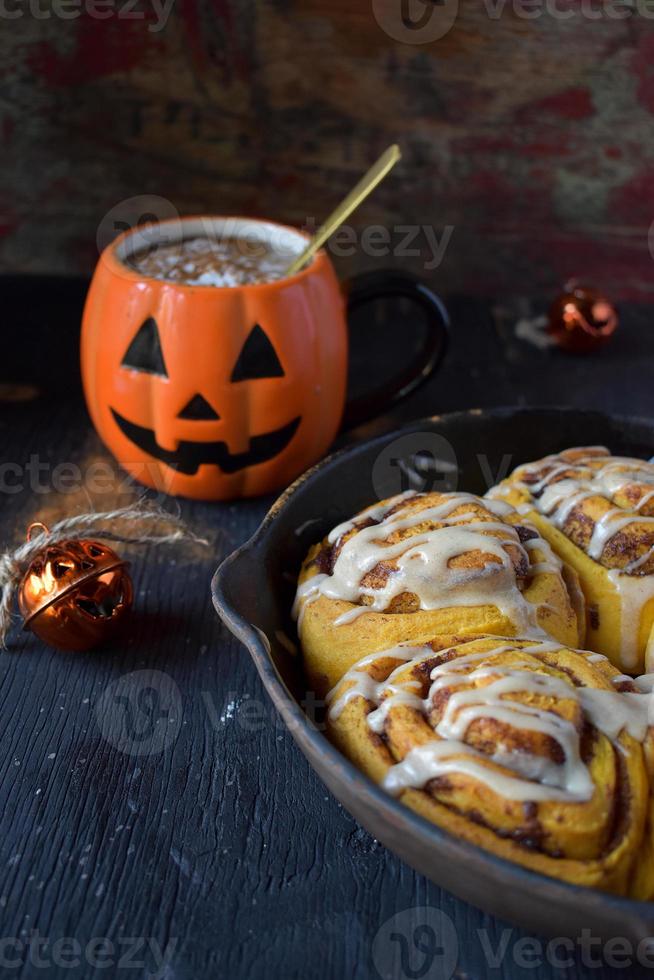 Cast iron pan of Halloween pumpkin cinnamon rolls drizzled with white icing with pumpkin mug drink photo