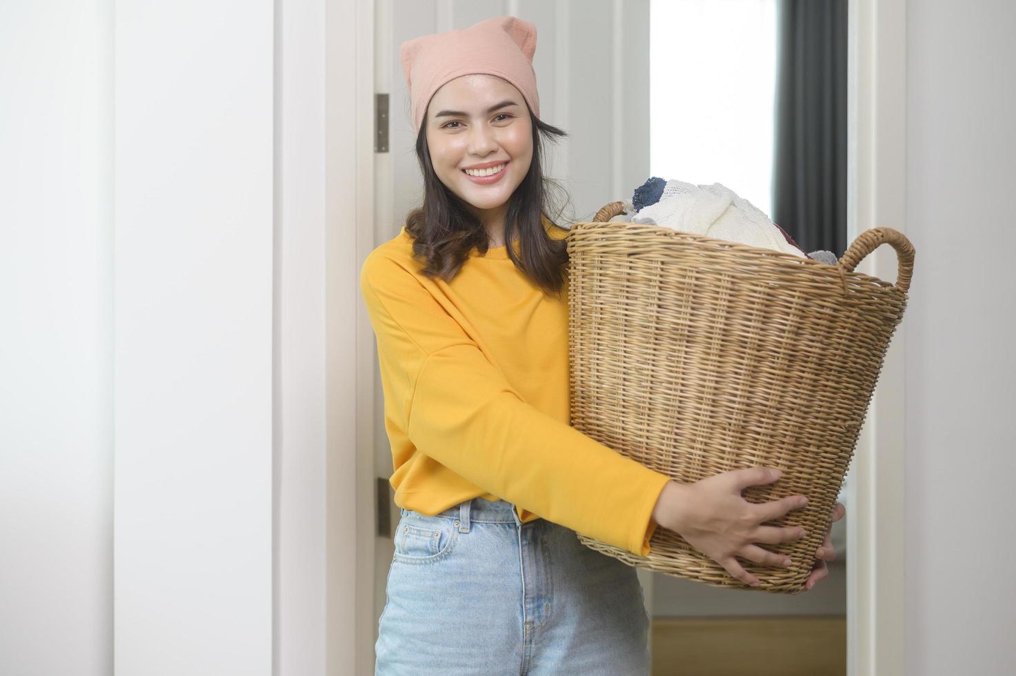 joven mujer feliz con pantalones amarillo sosteniendo una cesta llena de ropa en casa, concepto de lavandería foto