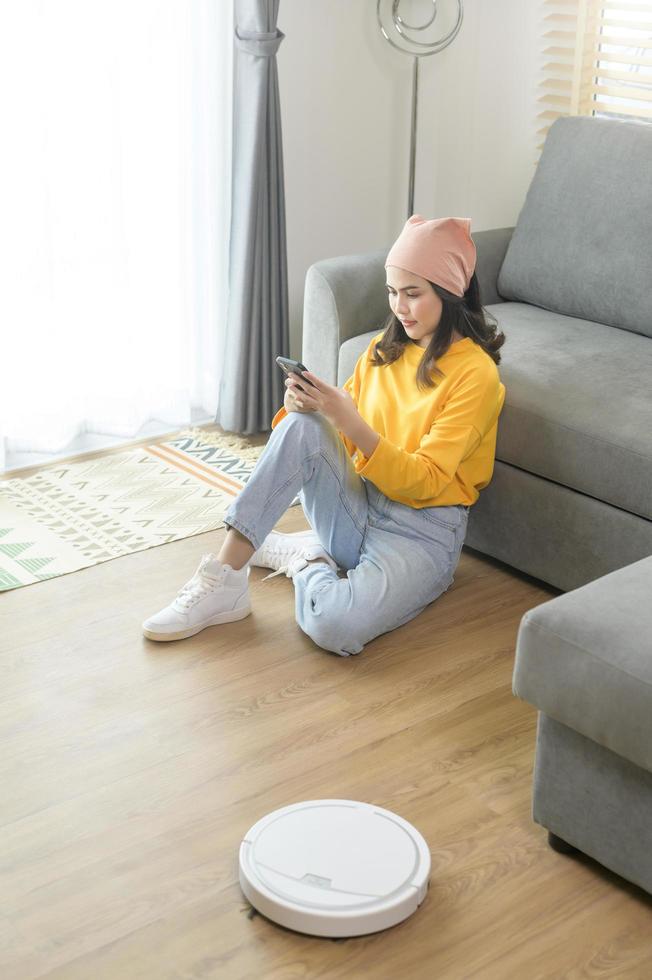 joven mujer feliz relajándose y usando un teléfono inteligente en la sala de estar mientras la aspiradora robótica funciona foto