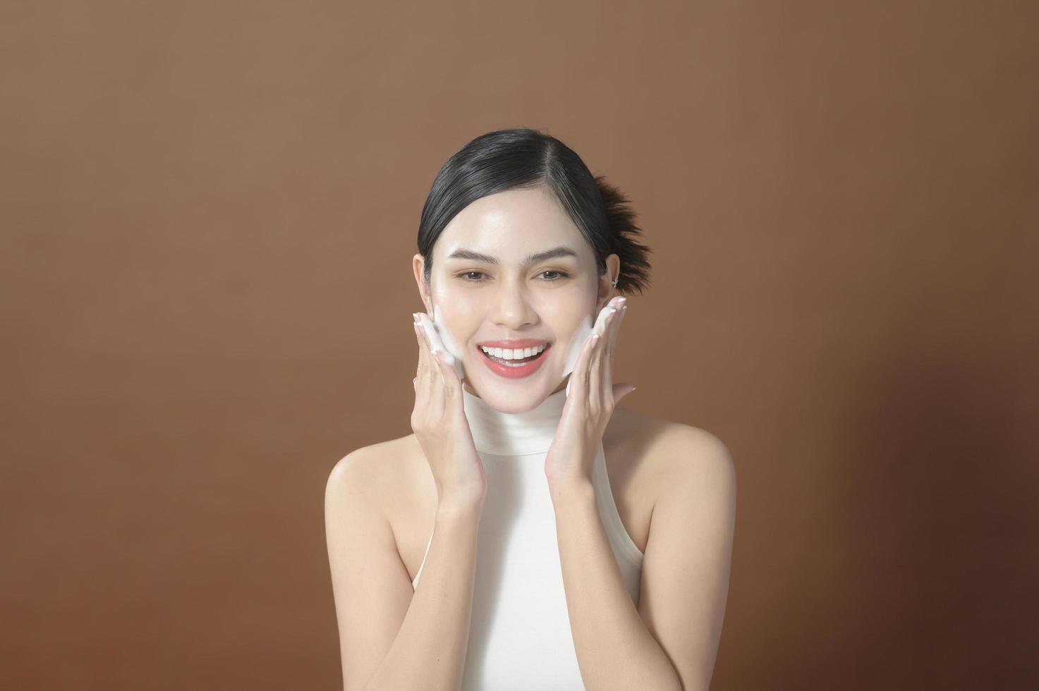 una mujer joven con cara hermosa sonriendo, lavándose la cara sobre fondo marrón, concepto de cuidado de la piel de belleza foto