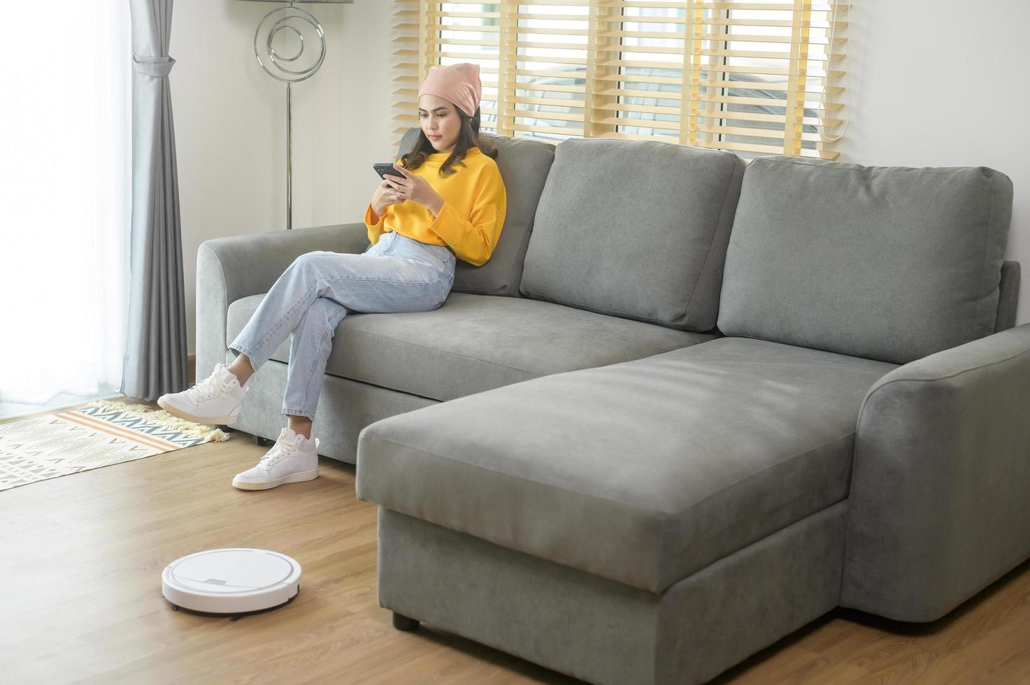 Young happy woman relaxing and using smartphone in living room while Robotic vacuum cleaner working photo