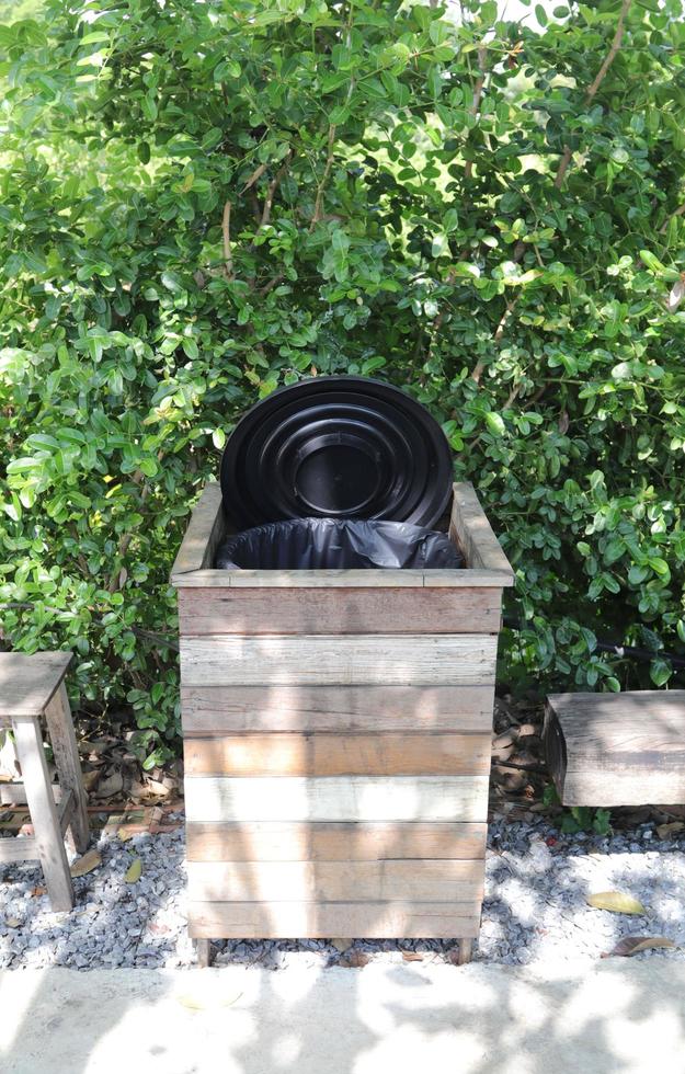 A vintage-style wooden trash can or bin in a park - a tourist attraction - with a visible black plastic bag inside, with a green tree background. photo