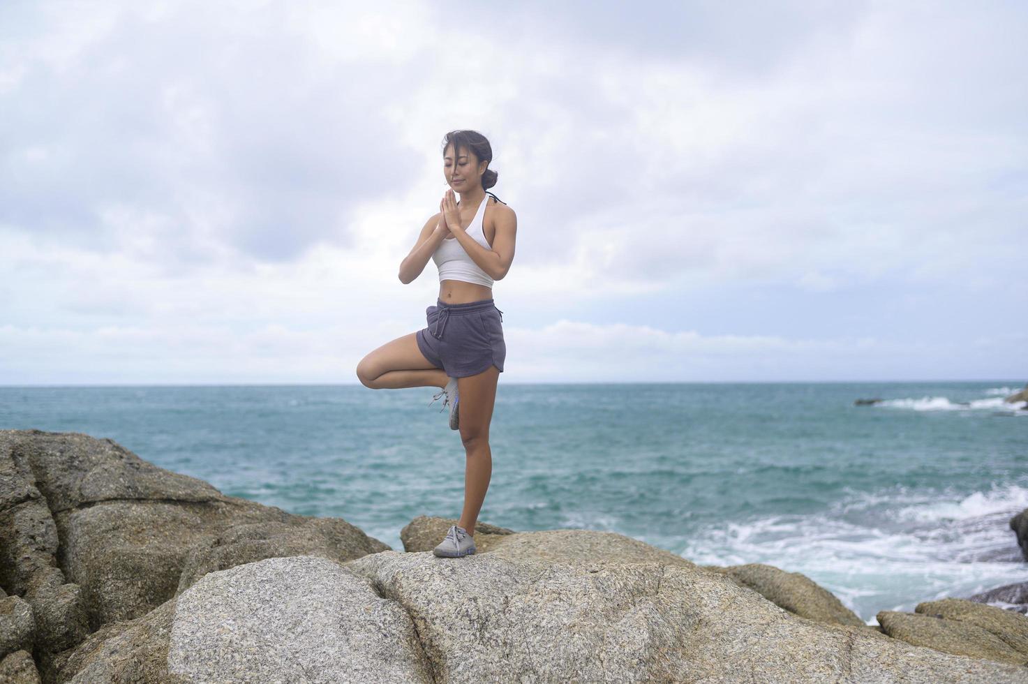 Young asian woman in sportswear doing yoga on the rock at seaside, health and meditation concept photo