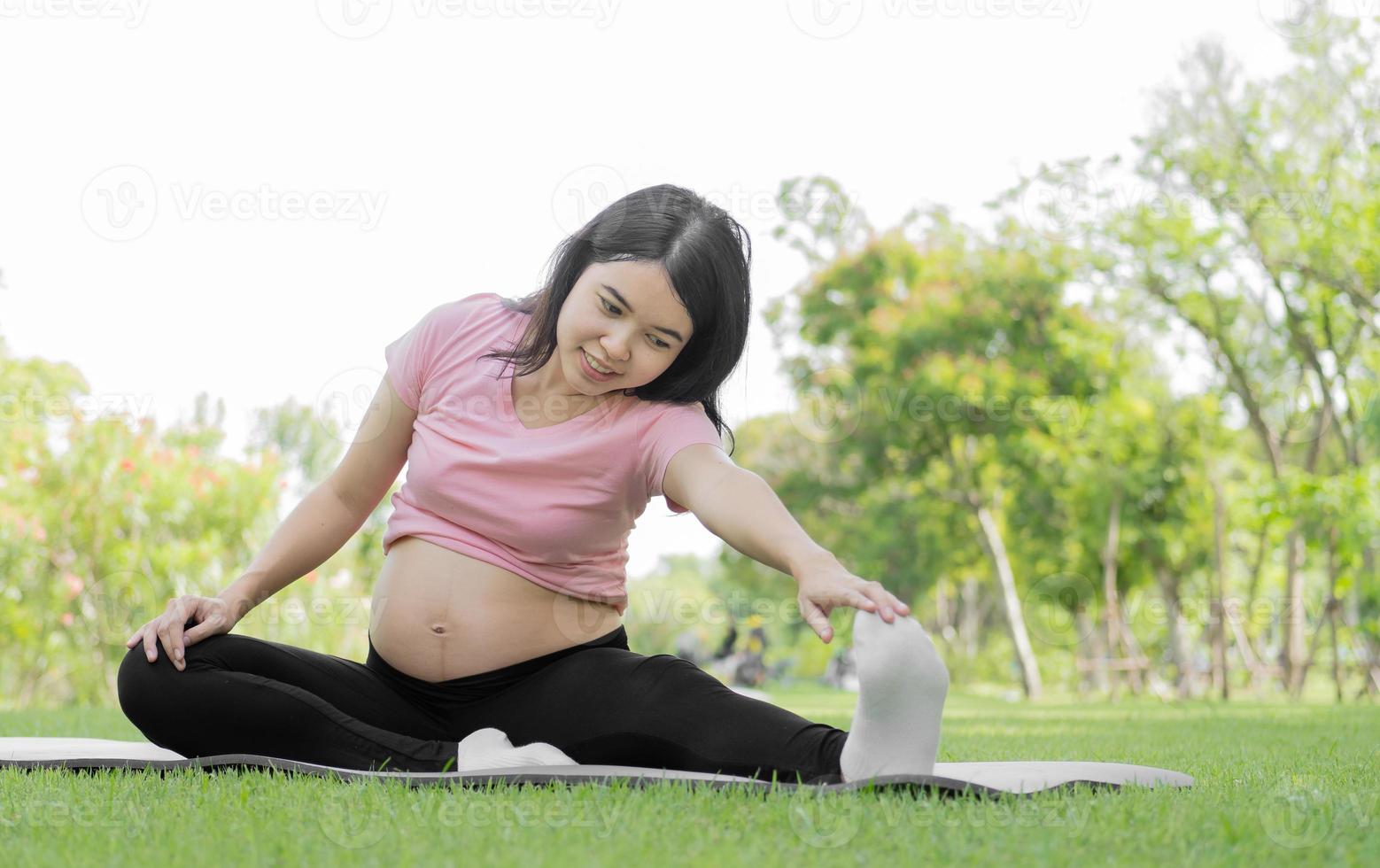 Happy pregnant woman sits relaxing outside in the park exercising. Young Asian pregnant woman is sitting on green grass in a nature garden doing yoga and stretching. Pregnancy and motherhood concept. photo