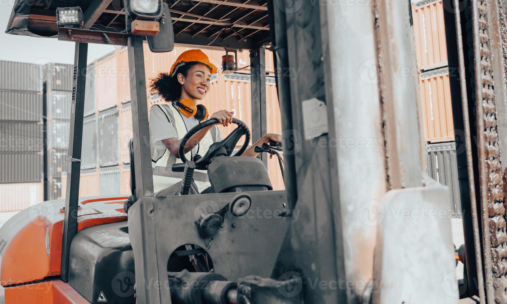 Female foreman with hard hat driving forklift at shipping container yard, portrait. Smiling multiracial industrial engineer woman in safety vest drives reach stacker to lift cargo box at logistic dock photo