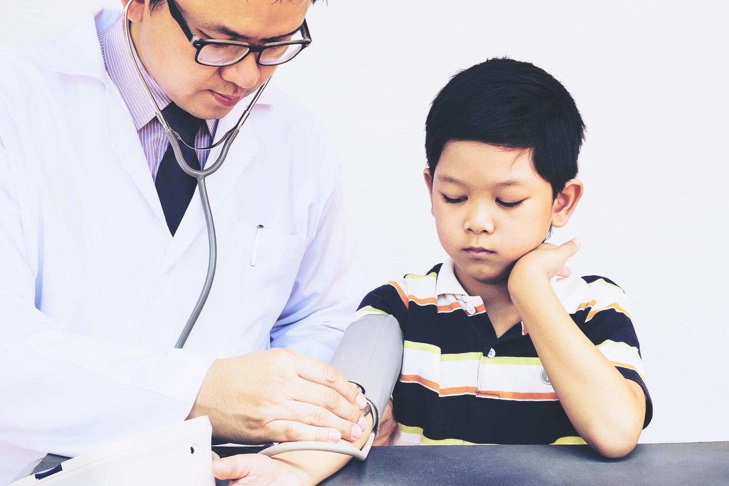 niño asiático siendo examinado por un médico varón usando estetoscopio y monitor de presión arterial sobre fondo blanco foto