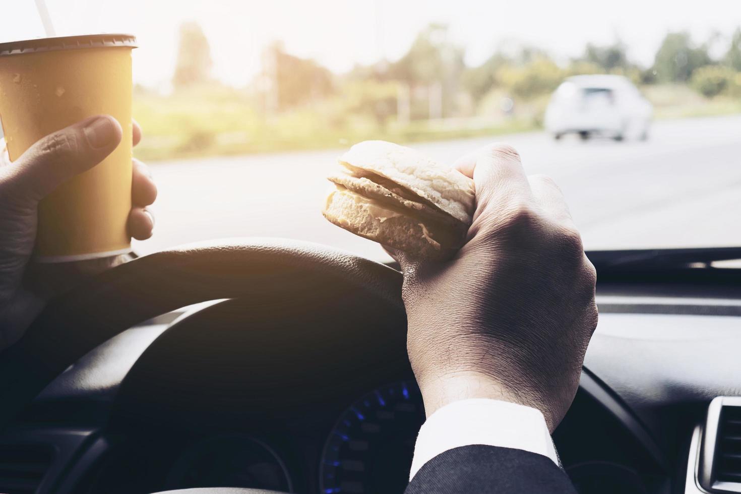 hombre conduciendo un auto mientras sostiene una taza de café frío y come una hamburguesa foto