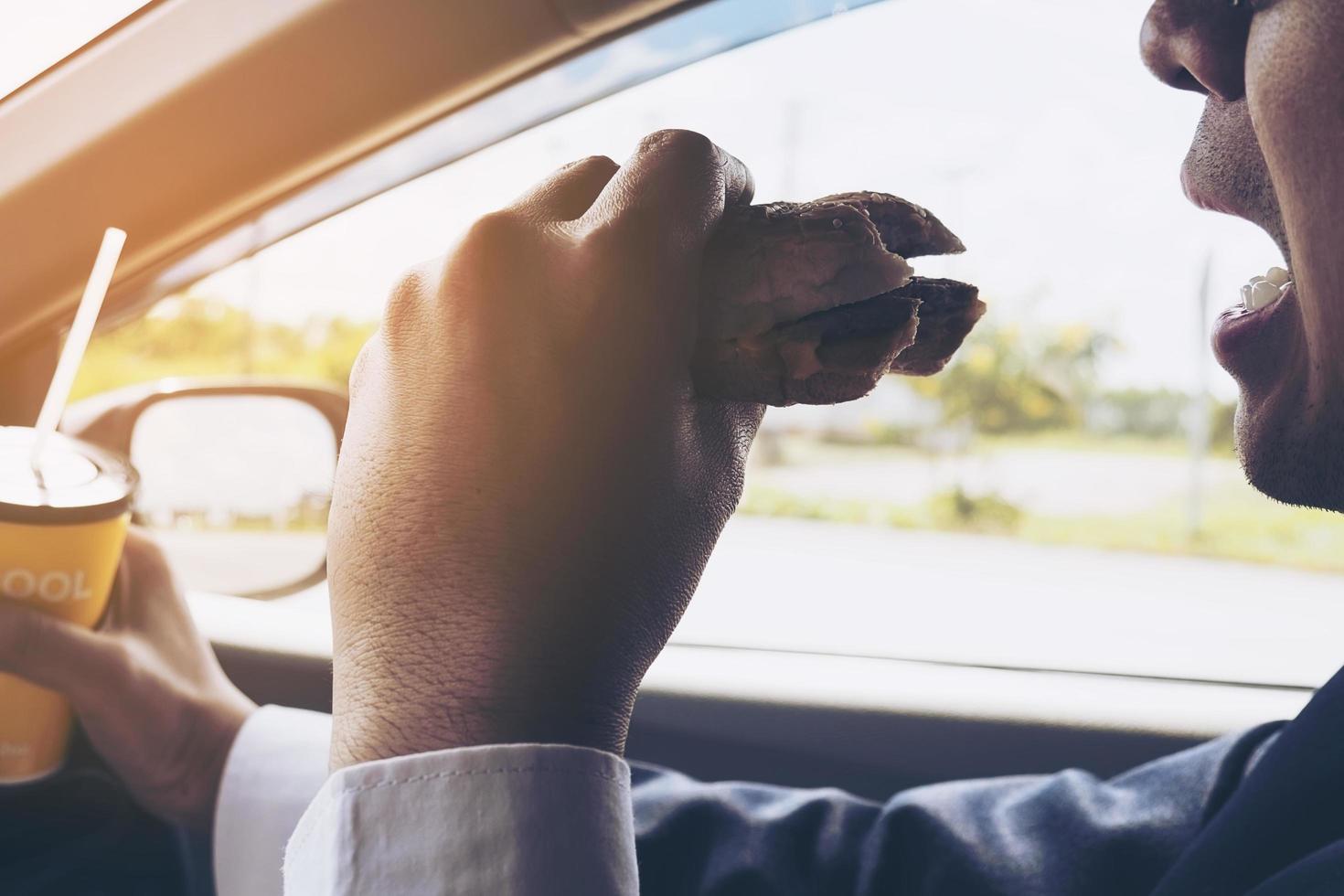 hombre conduciendo un auto mientras sostiene una taza de café frío y come una hamburguesa foto