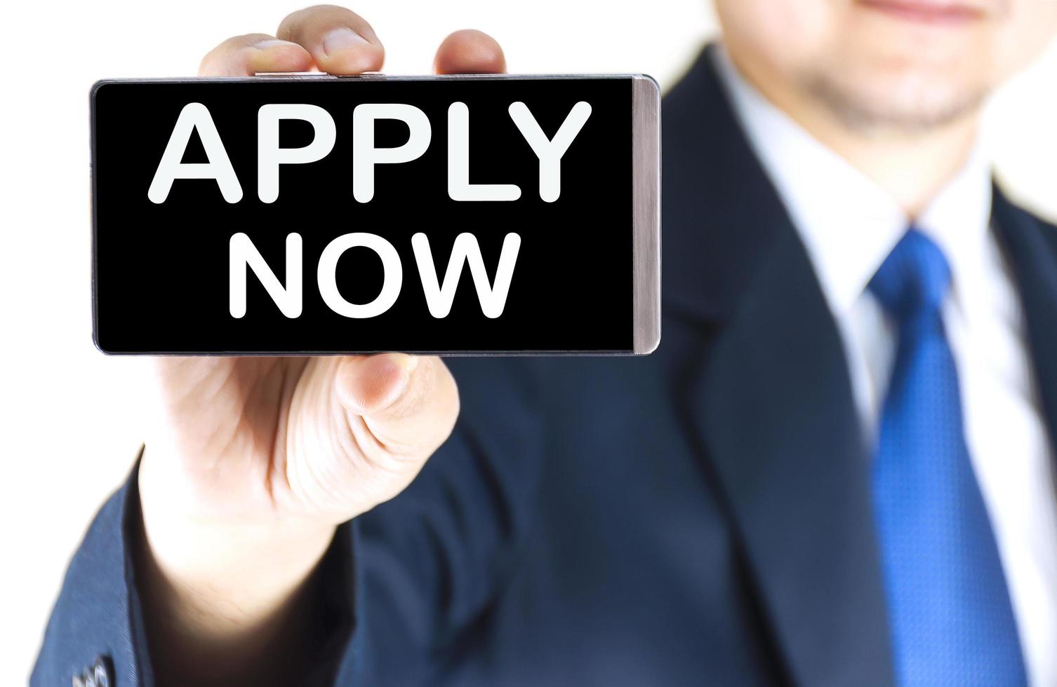 APPLY NOW, word on mobile phone screen in blurred young businessman hand over white background, business concept photo