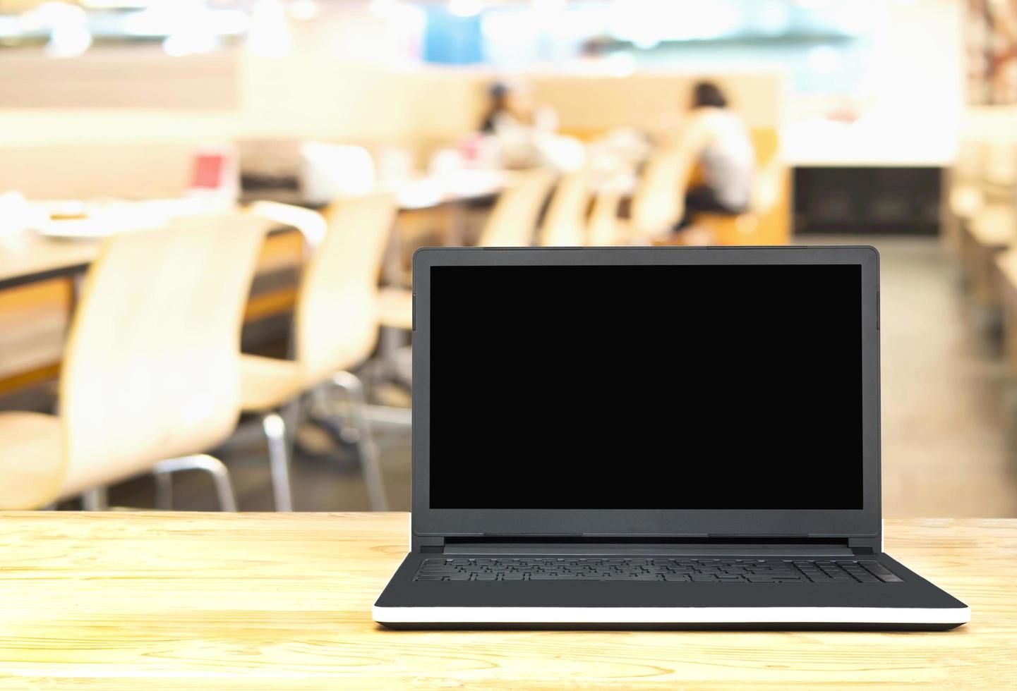 Blank screen laptop on wooden table with blurred people in restaurant background. Photo includes CLIPPING PATH of blank screen.