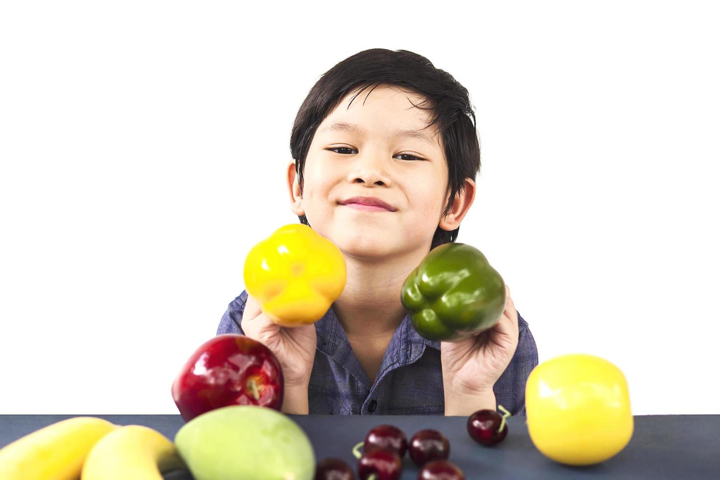 Chico sano asiático mostrando expresión feliz con variedad de frutas y verduras de colores sobre fondo blanco. foto