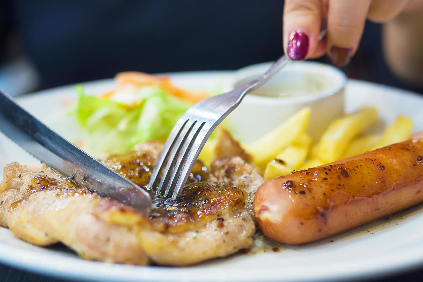 primer plano de personas dejando caer salsa al filete de pollo con salchichas, papas fritas y plato de ensalada foto