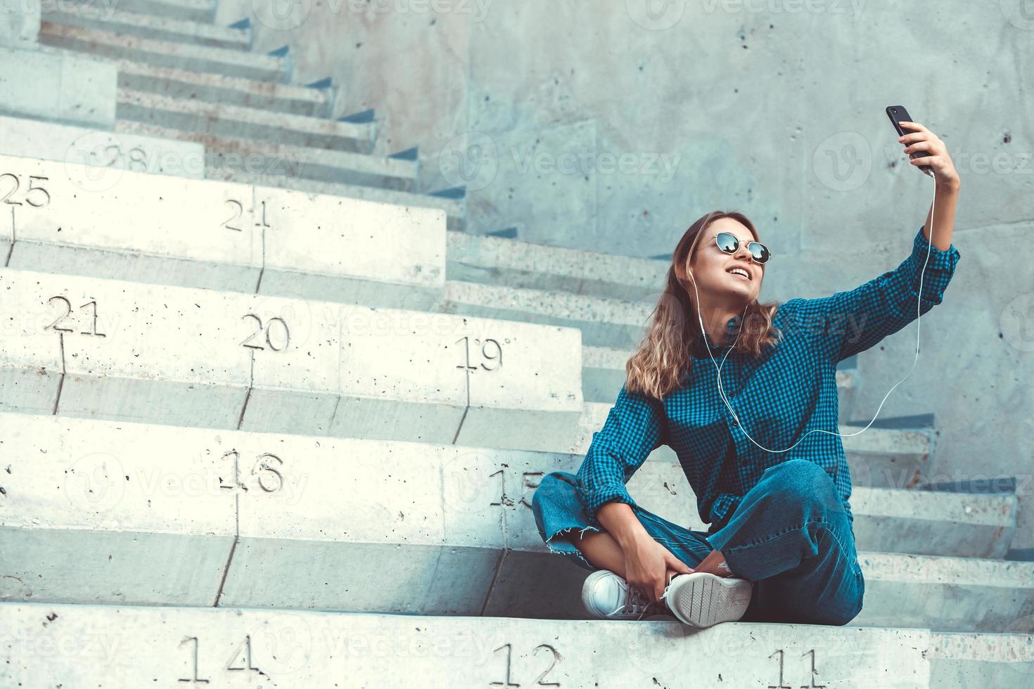 foto de una alegre y linda joven estudiante sonriente con gafas de sol al aire libre usando un teléfono móvil charlando escuchando música con auriculares.