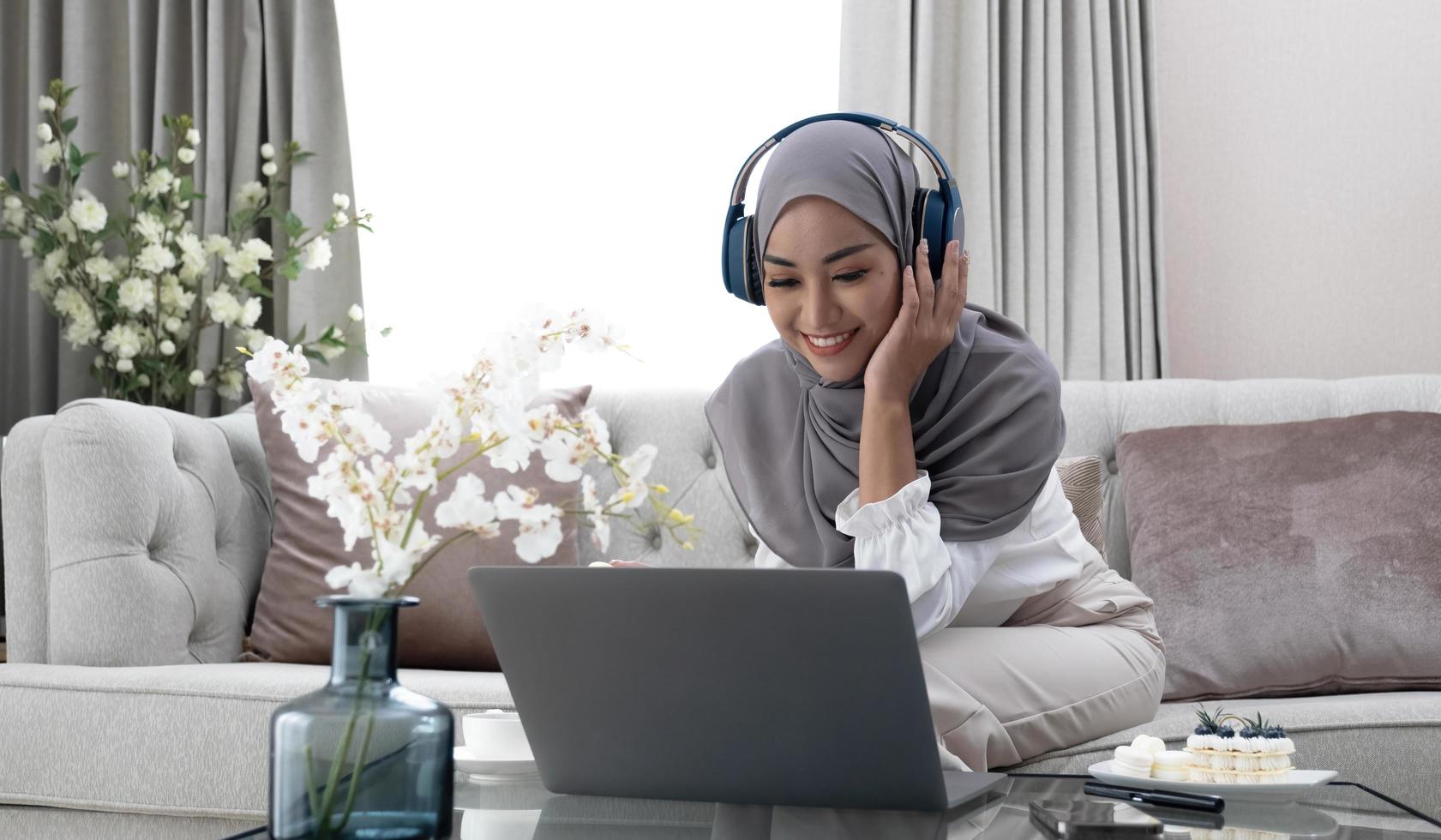 lista de reproducción para relajarse retrato de una mujer musulmana tranquila en hijab escuchando música con su computadora portátil y auriculares inalámbricos en casa. una mujer islámica sonriente se sienta con los ojos cerrados foto