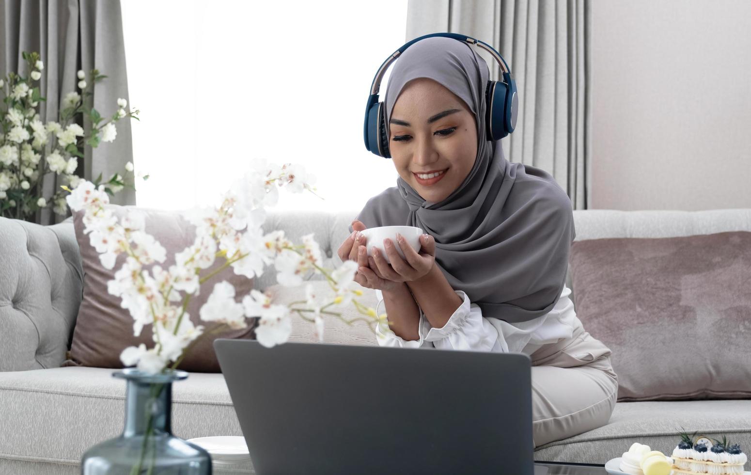 estilo de vida doméstico. mujer musulmana feliz usando una laptop en casa, viendo películas y tomando café, relajándose en el sofá en la sala de estar, vista lateral foto