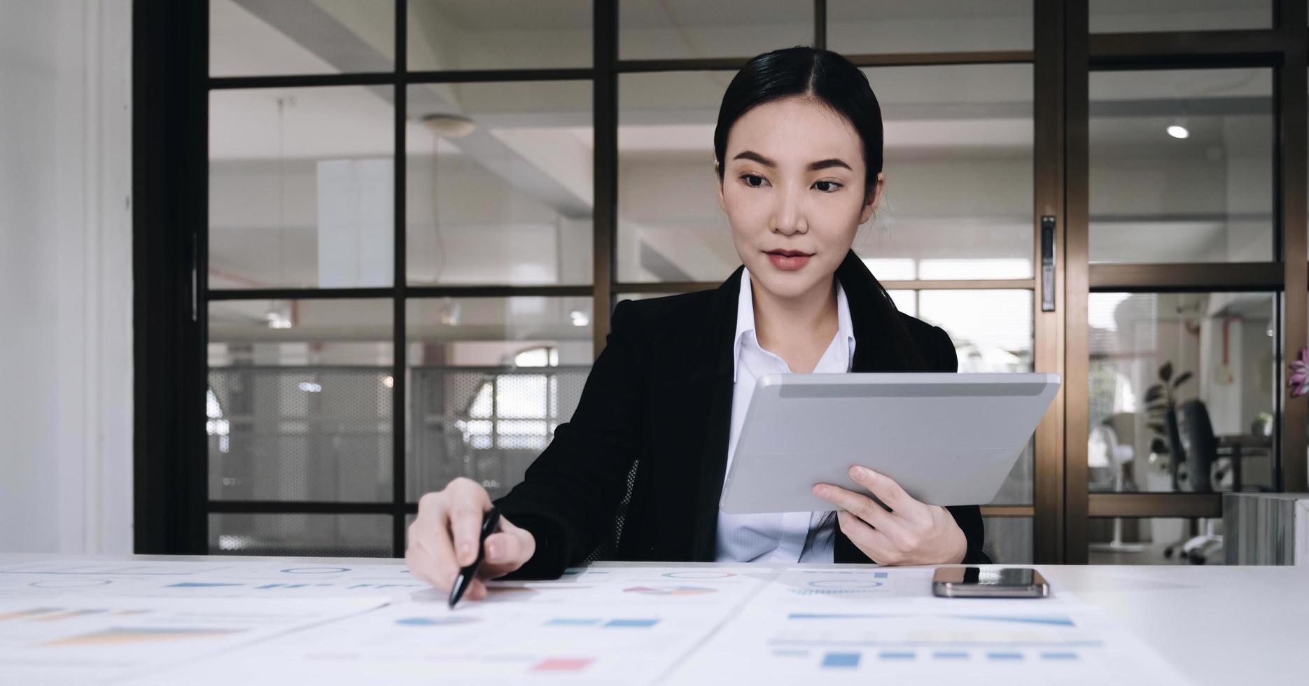 Attractive and charming young Asian businesswoman or financial worker in formal suit using tablet and working with her financial report in the office. photo