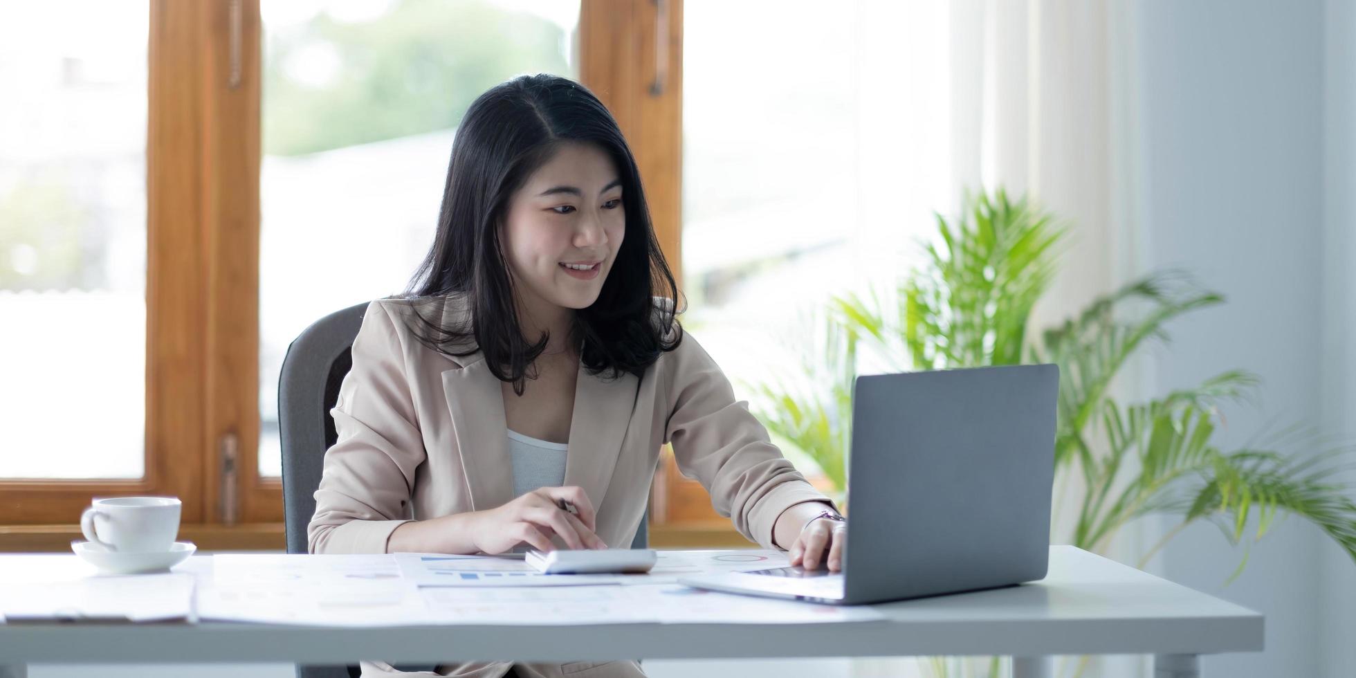 Asian businesswoman working in financial accounting analyzes a chart using a laptop calculator with documents and holding a pen sitting on a chair at an office desk photo