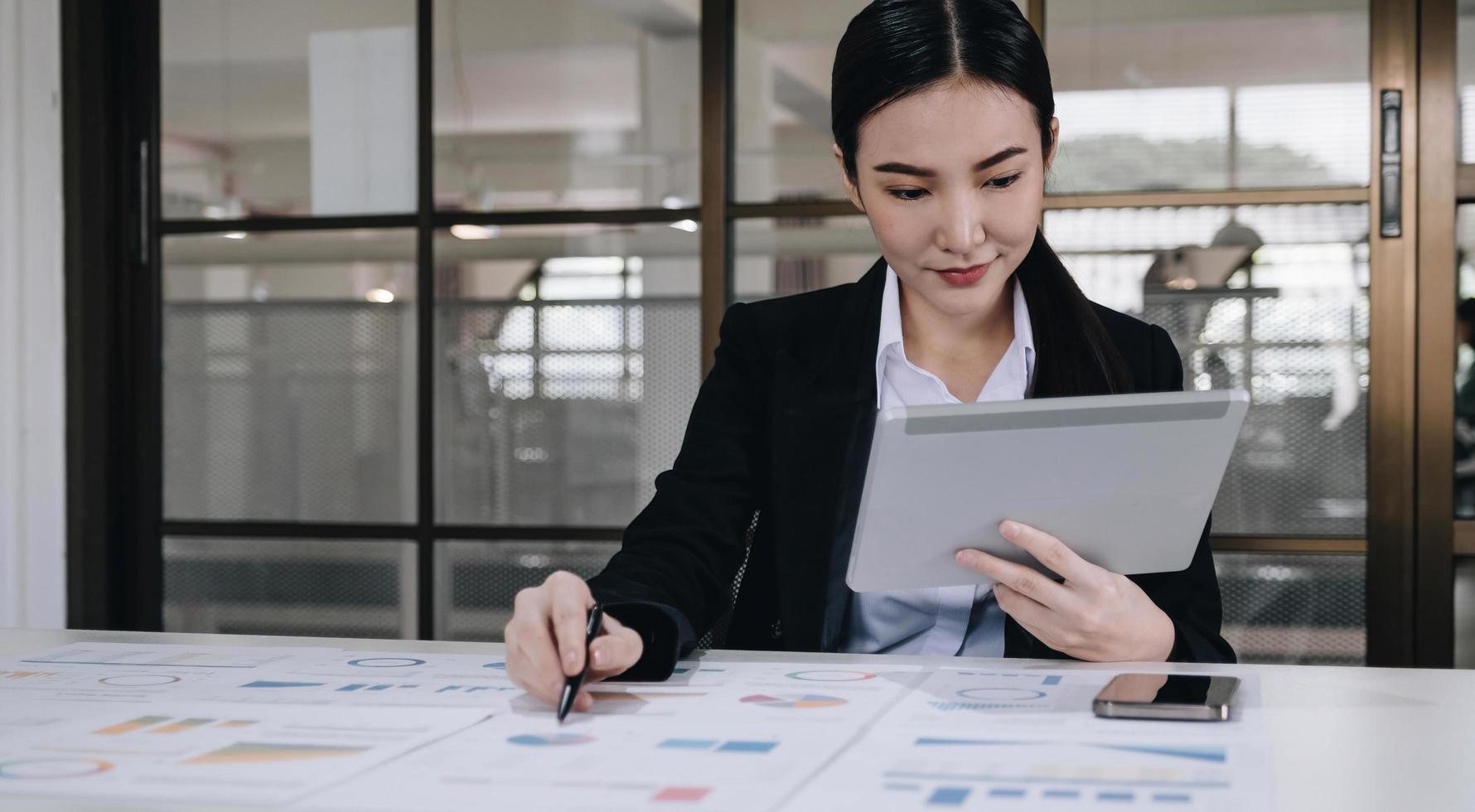 Attractive and charming young Asian businesswoman or financial worker in formal suit using tablet and working with her financial report in the office. photo