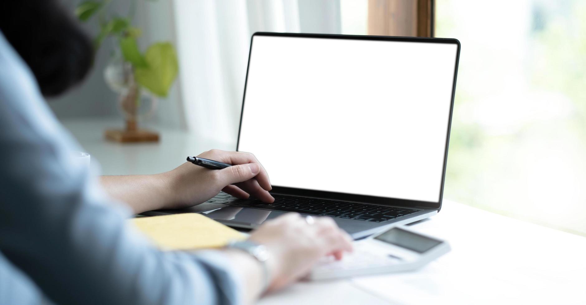 Mockup image of a woman using laptop with blank white screen on wooden table in office photo