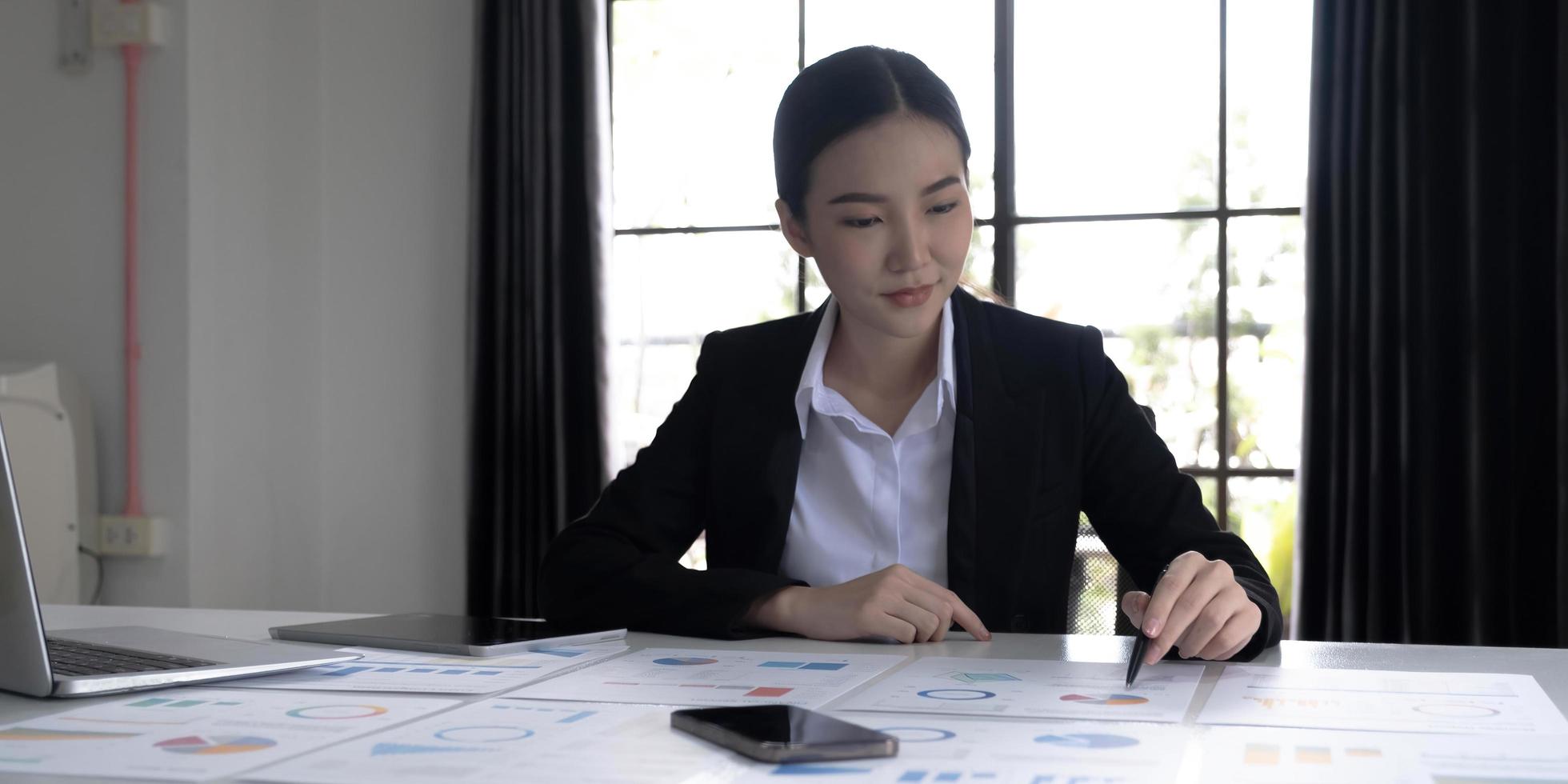 Young Asian businesswoman analyzing report pointing at graph holding a pen using a tablet laptop placed at the office. photo