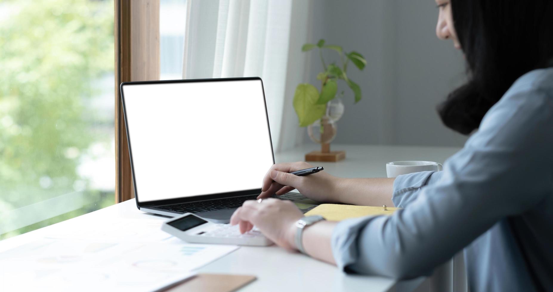 Mockup image of a woman using laptop with blank white screen on wooden table in office photo