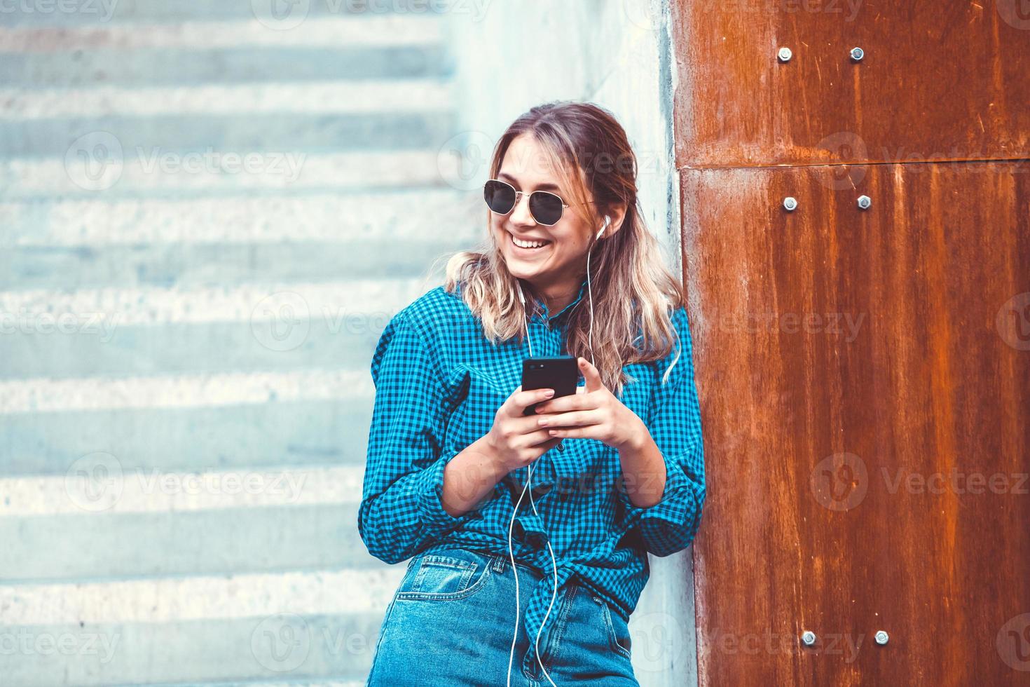 Photo of a cheerful smiling cute young student girl wearing sunglasses outdoors using mobile phone chatting listening music with earphones.