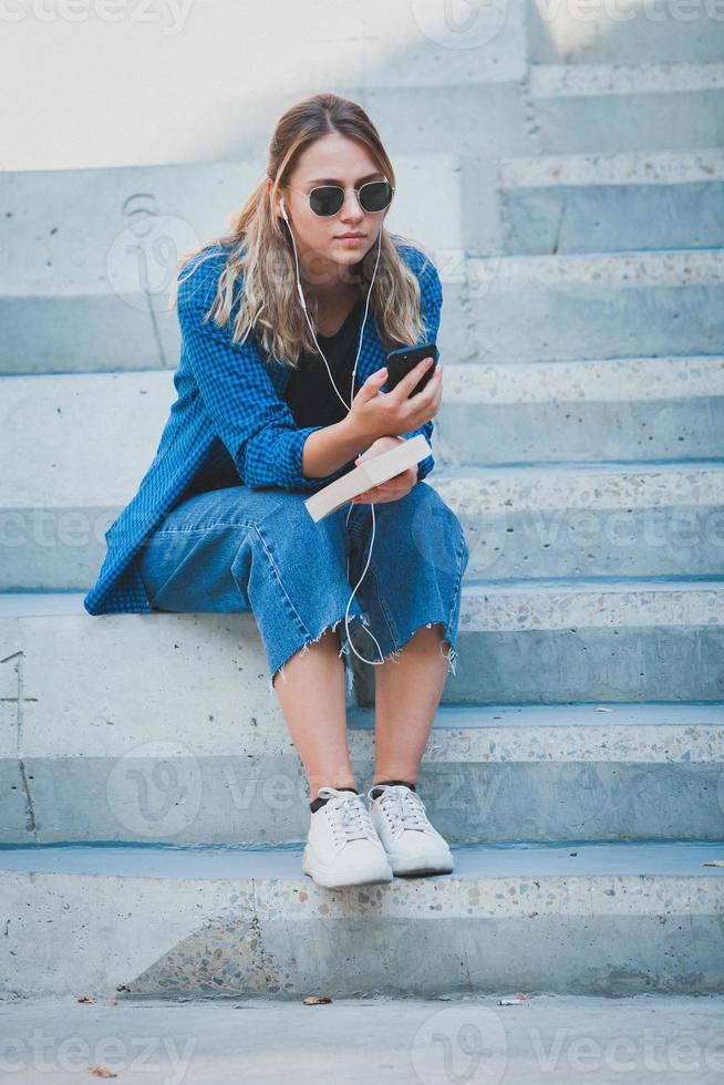 Photo of a cheerful smiling cute young student girl wearing sunglasses outdoors using mobile phone chatting listening music with earphones.