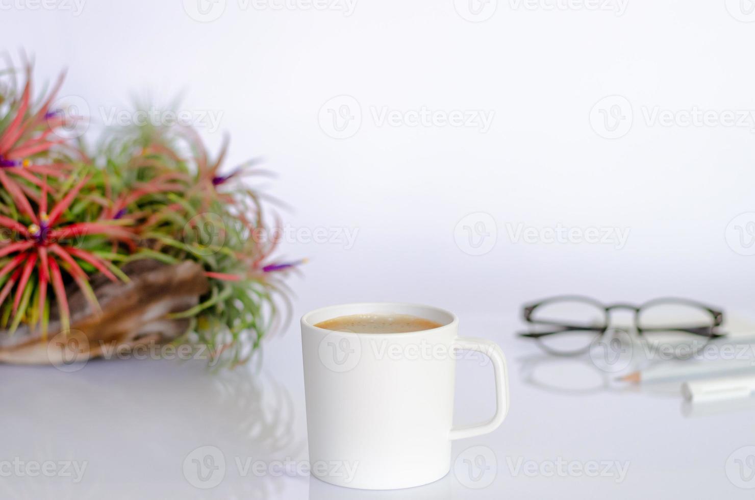 A cup of coffee on working table with air plant Tillandsia, spectacles, pen and pencil on white background. photo