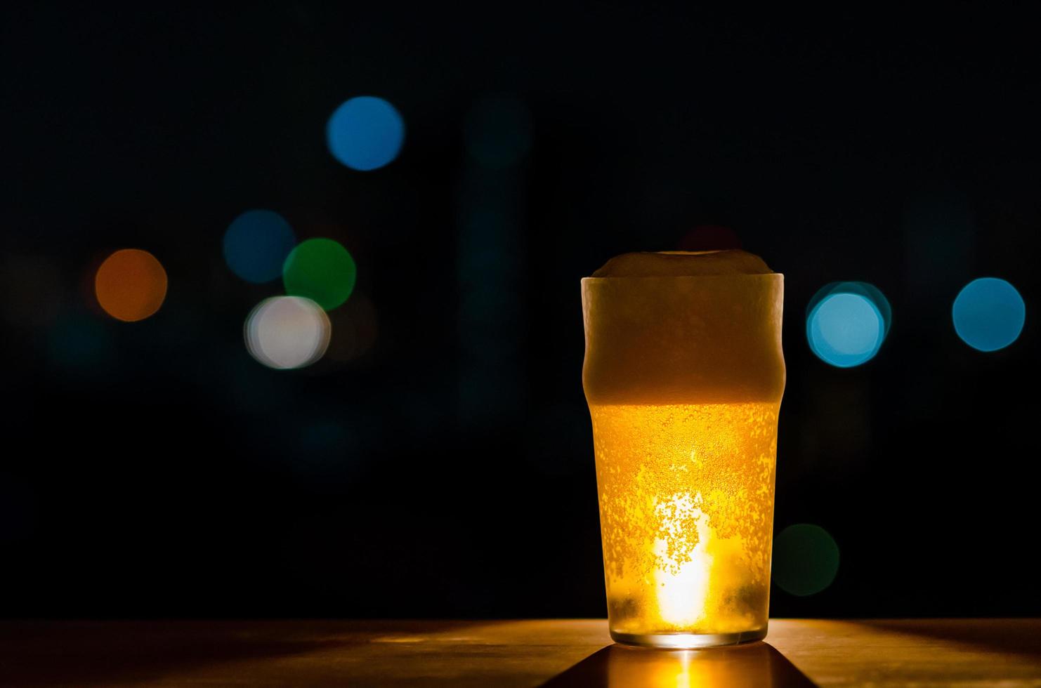 A glass of Beer with its foam puts on wooden table of the bar isolated on dark night background with colorful bokeh lights on rooftop bar. photo