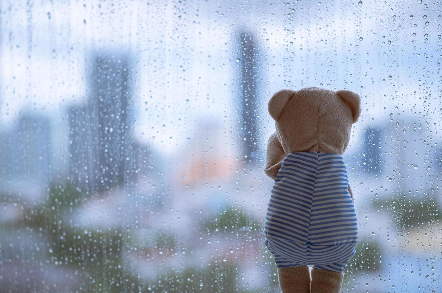 Teddy bear crying alone at window when raining with blurred city background. photo