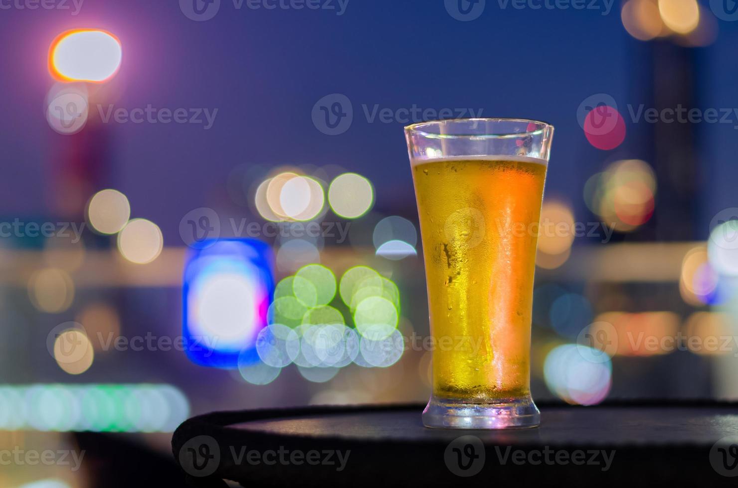 A glass of beer puts on table at rooftop bar with colorful city bokeh light background. photo