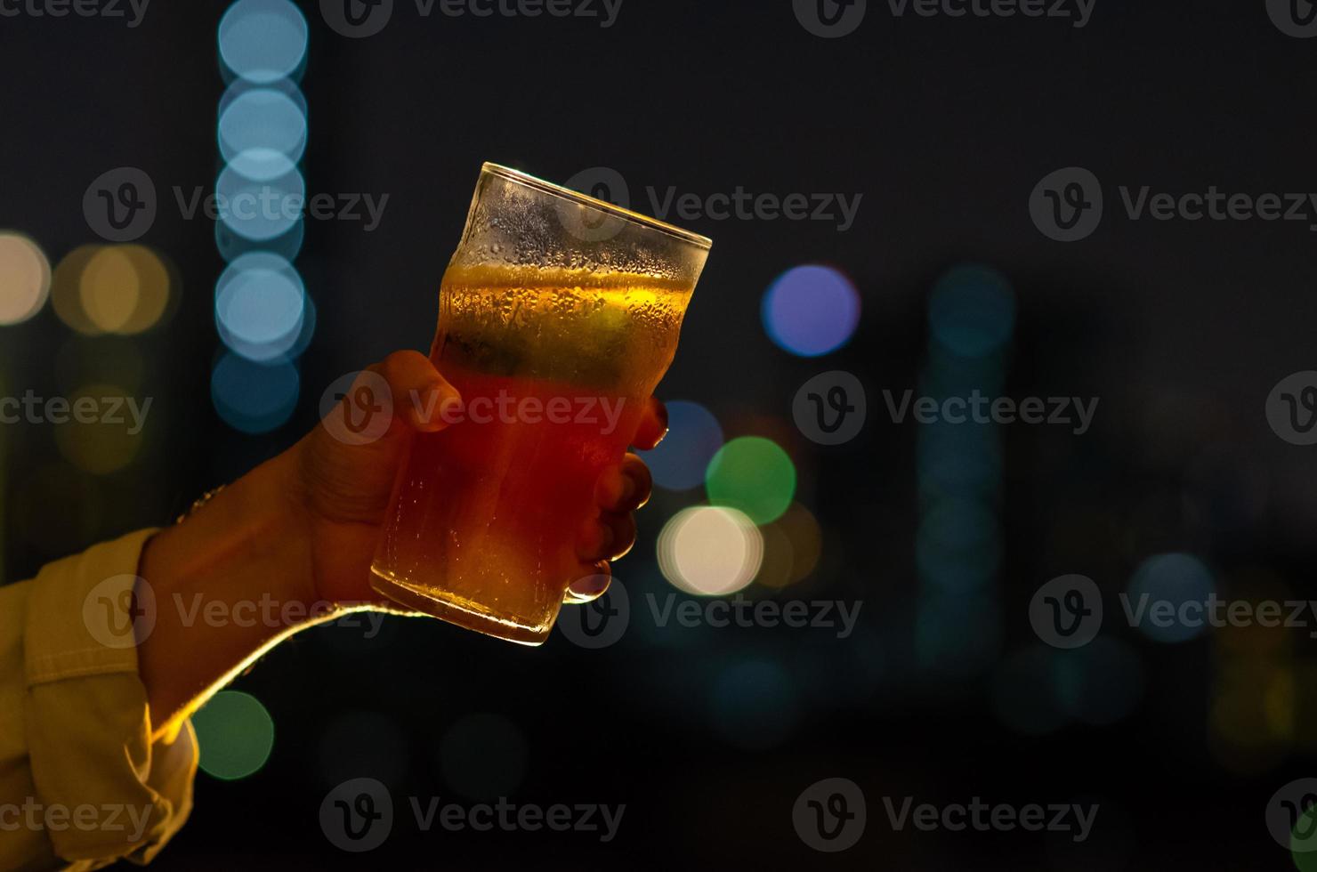 Hand with glass of beer toasting for celebration and party concept isolated on dark night background with colorful city bokeh lights on rooftop bar. photo