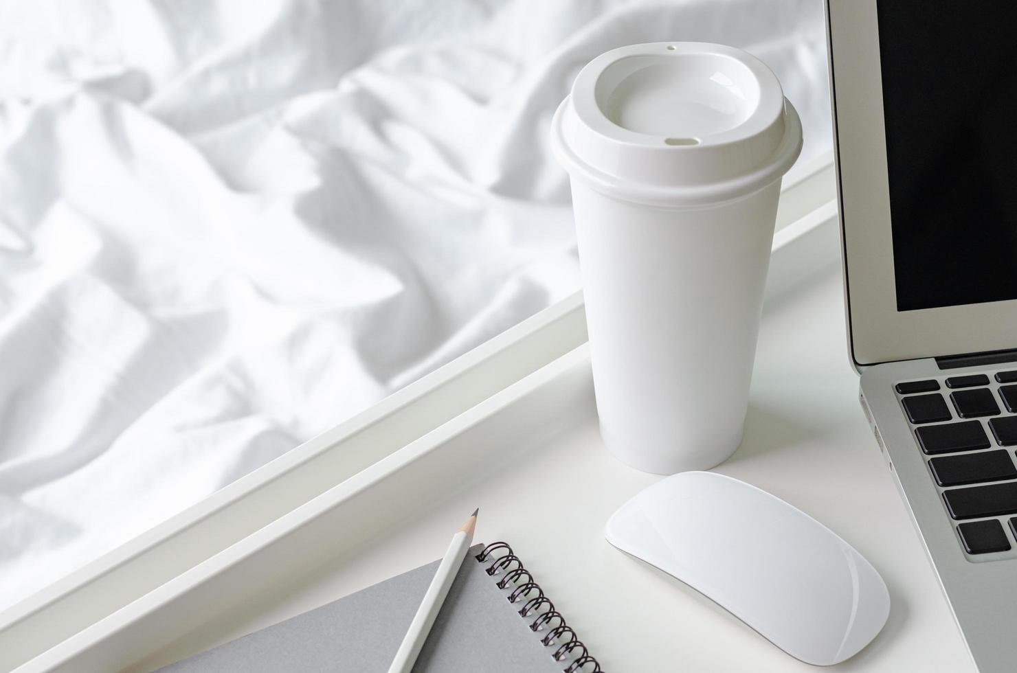 una taza de café se coloca al lado de la computadora portátil y el mouse con una bandeja blanca sobre una manta desordenada para trabajar en el concepto de cama. foto