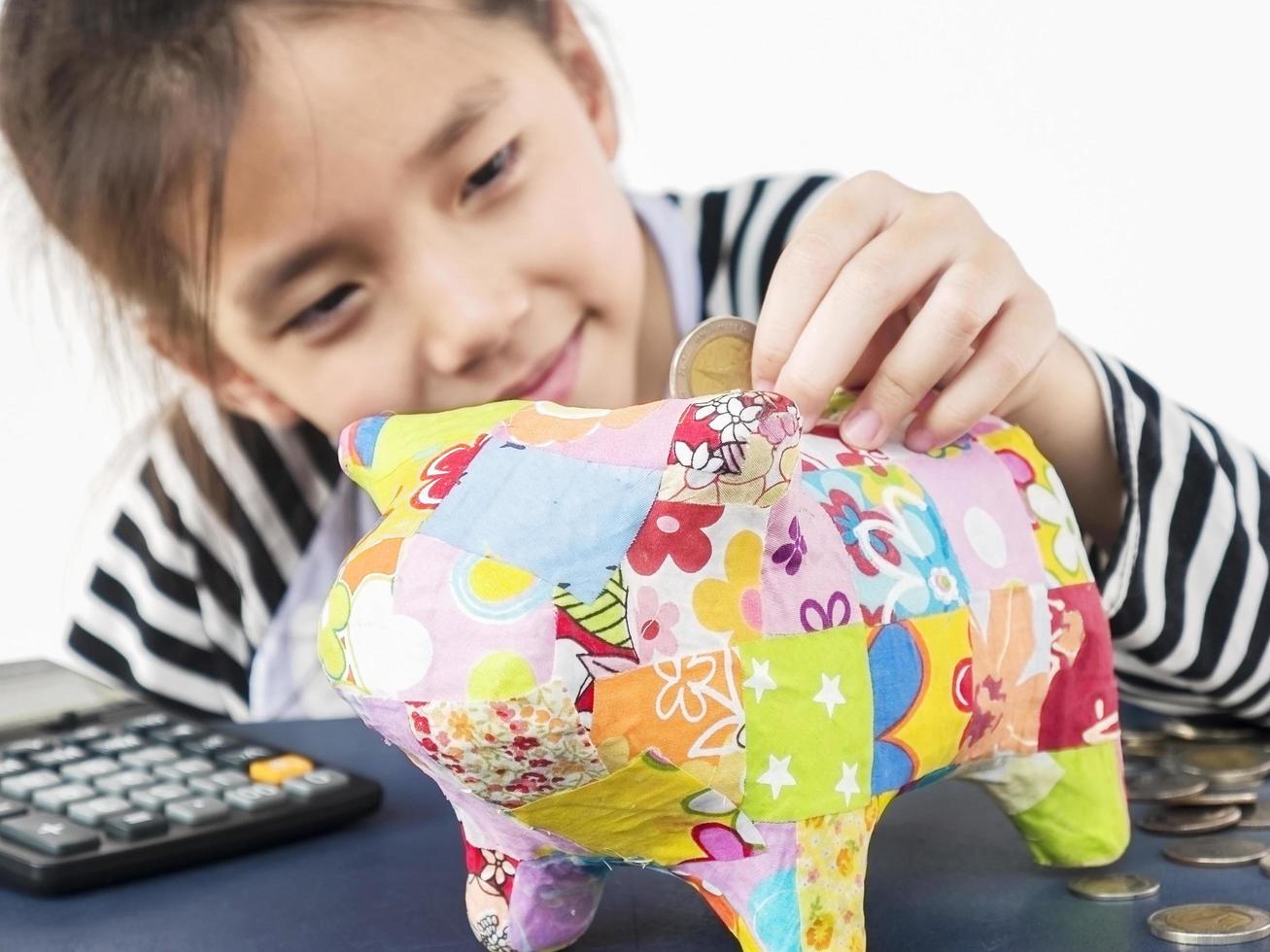 un niño asiático está poniendo dinero en un colorido banco de monedas con una calculadora en una mesa de madera foto