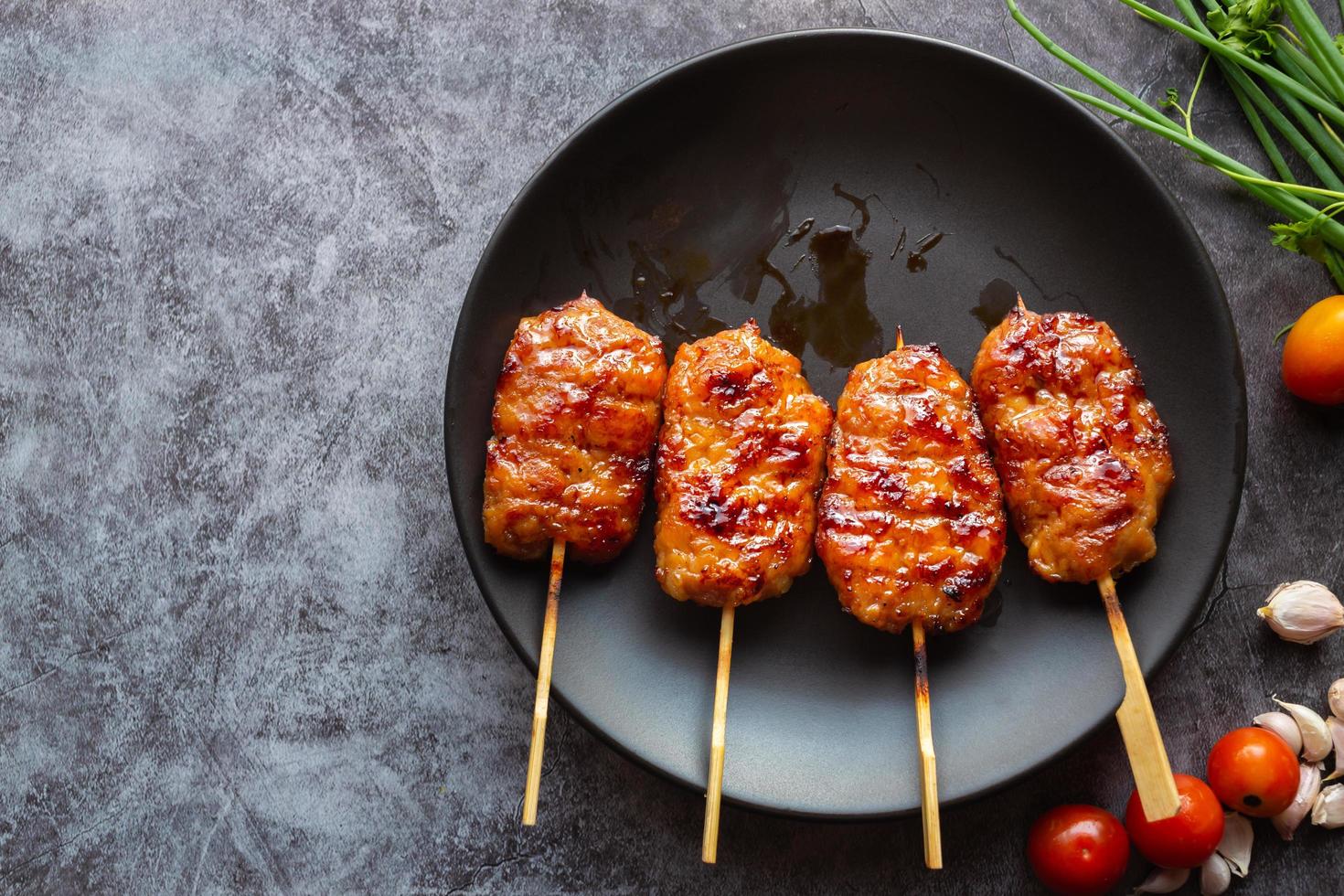 Grill skewered milk pork in a black dish with tomatoes with garlic and spring onion on a gray concrete background. Thai street food.Top view and copy space photo