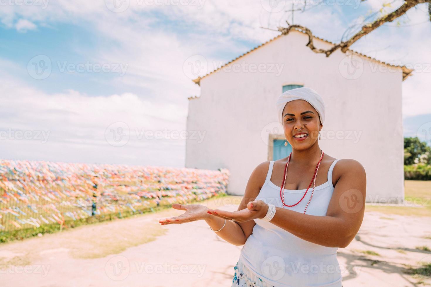 Happy Brazilian woman dressed in traditional Bahian costume showing something on the palm of her hand, looks at camera, with Porto Seguro historic center in the background photo