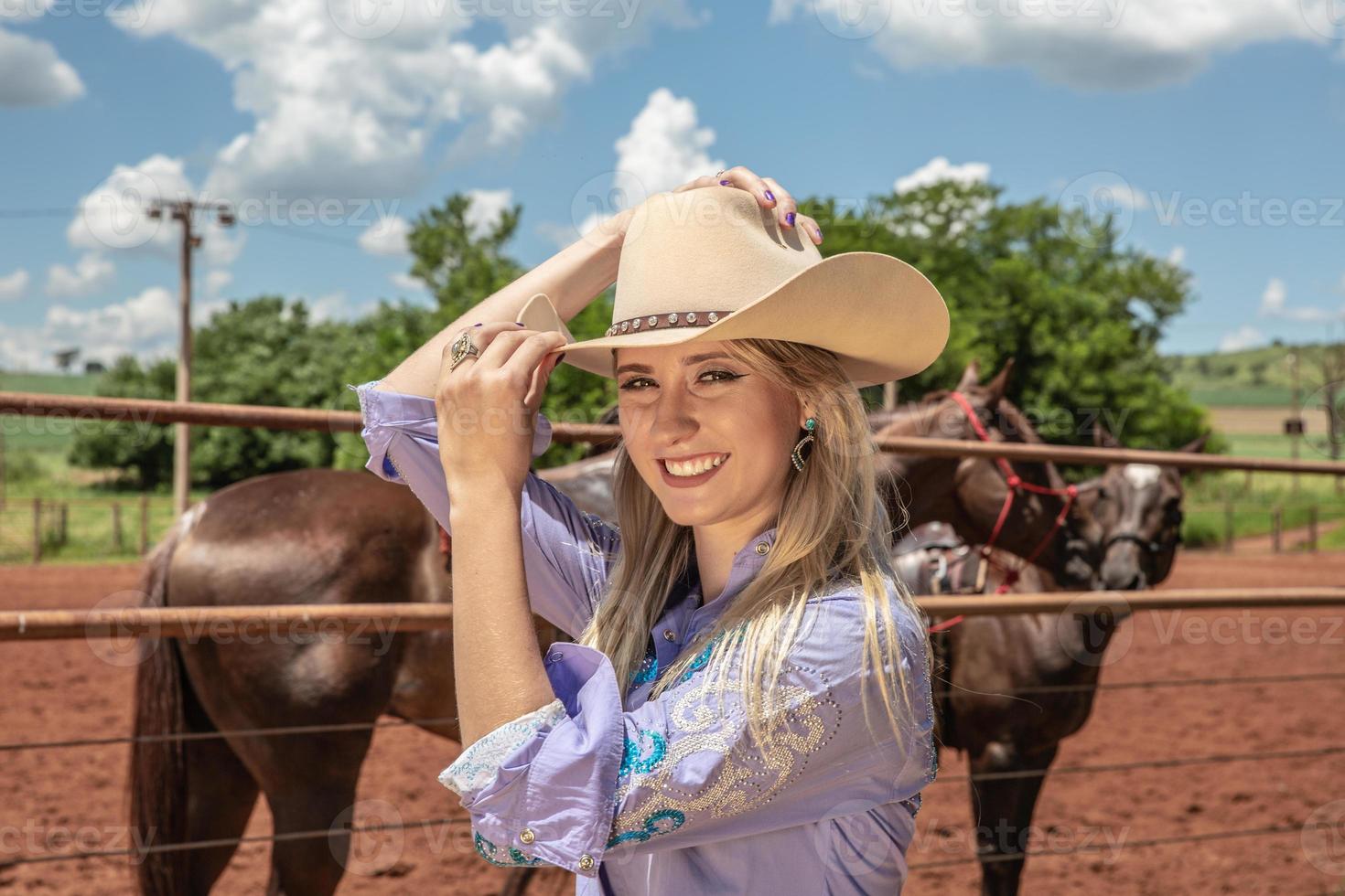 hermosa vaquera rubia con sombrero de pie cerca del fondo del rancho de caballos foto