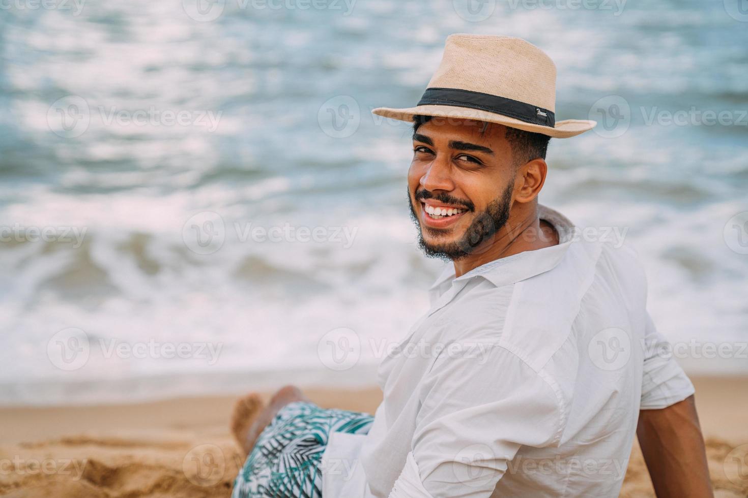 silueta de un joven en la playa. hombre latinoamericano sentado en la arena de la playa, con un sombrero, mirando a la cámara. un hermoso día de verano foto