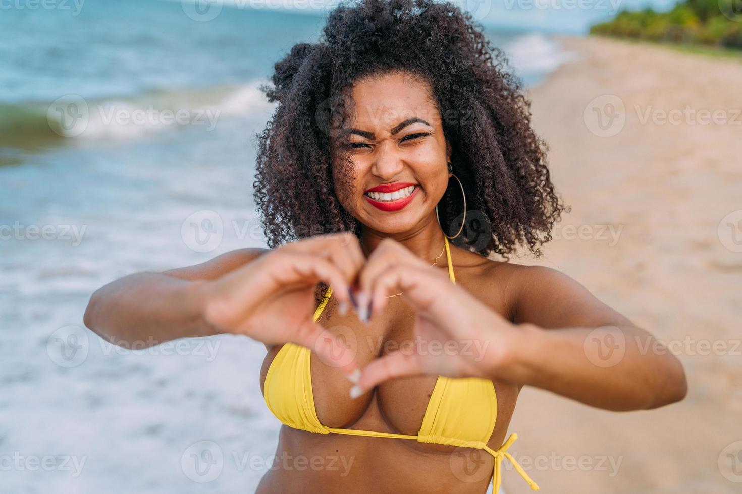 beautiful latin american woman in bikini on the beach. Young woman enjoying  her summer vacation on a sunny day, smiling, making a heart with her hands  and looking at the camera 9348904