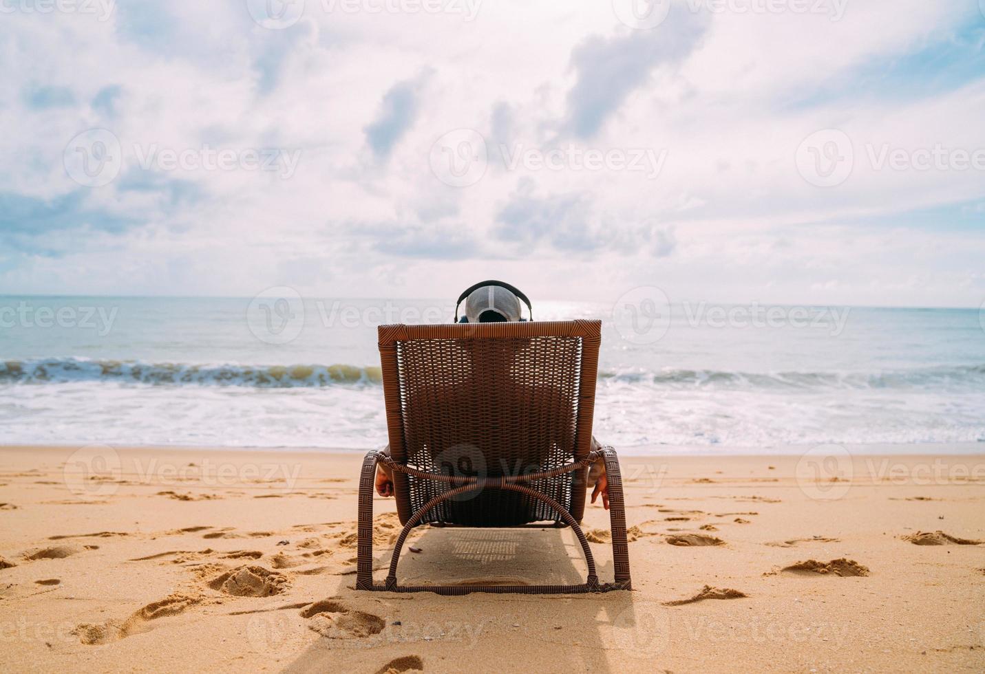 summer holidays, technology and internet concept. latin american man sitting on the beach chair, listen music with headphone and sunning on the beach photo
