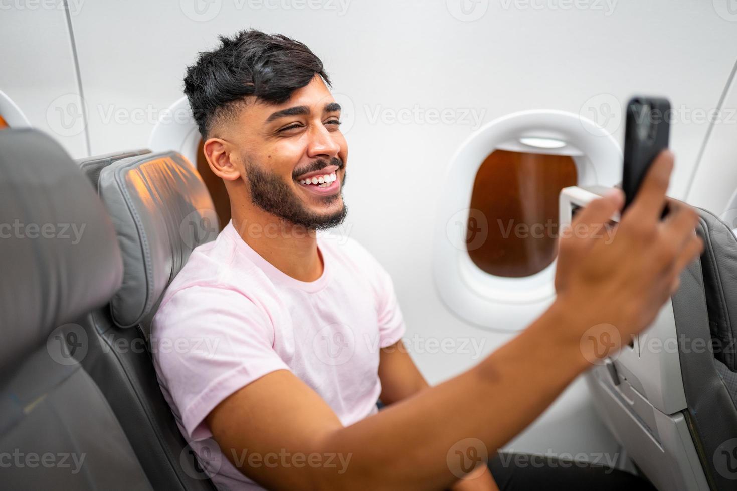 Young latin american man smiling making a video call on his cell phone, sitting on the plane near the window. photo