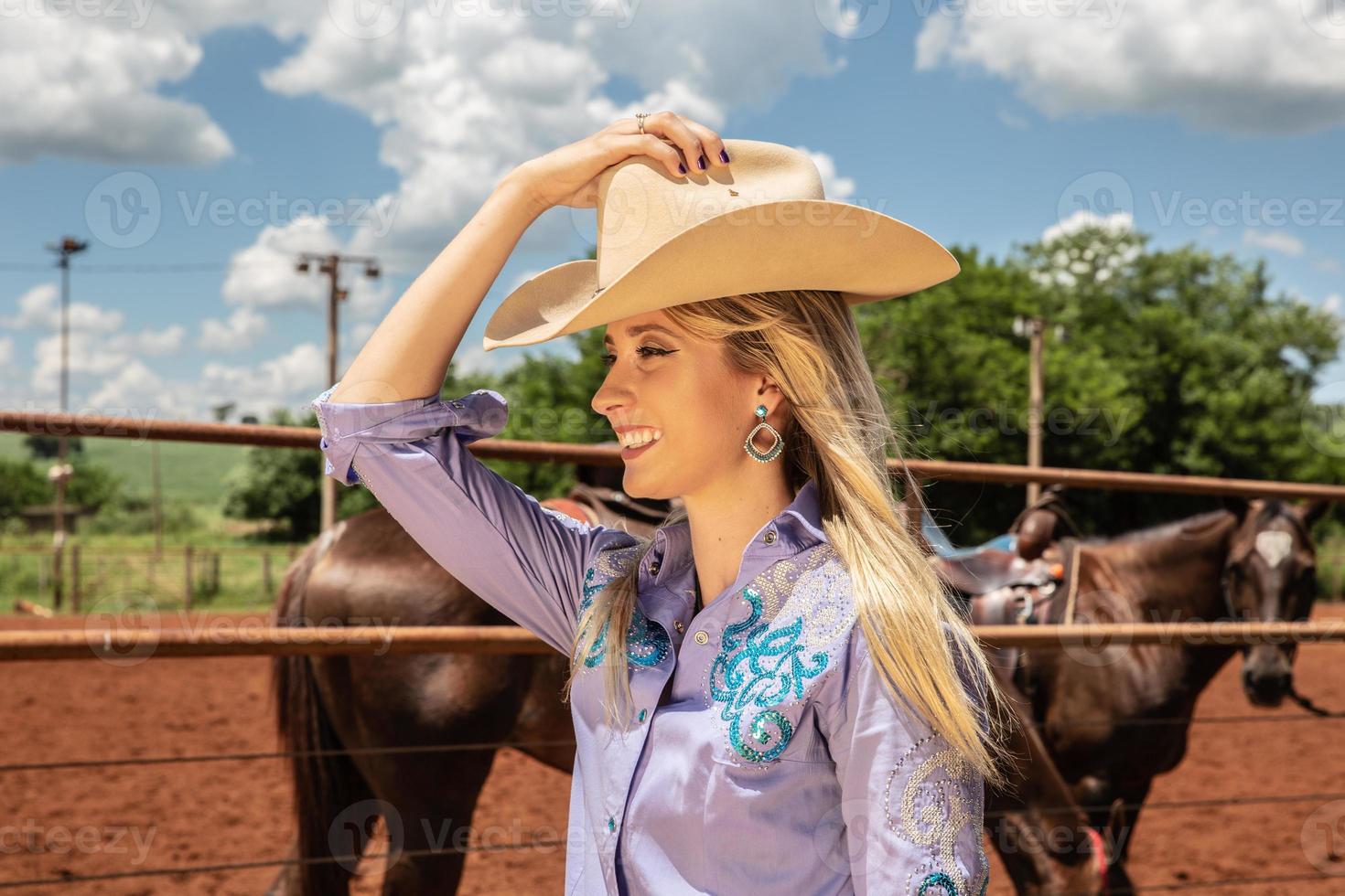 hermosa vaquera rubia con sombrero de pie cerca del fondo del rancho de caballos foto