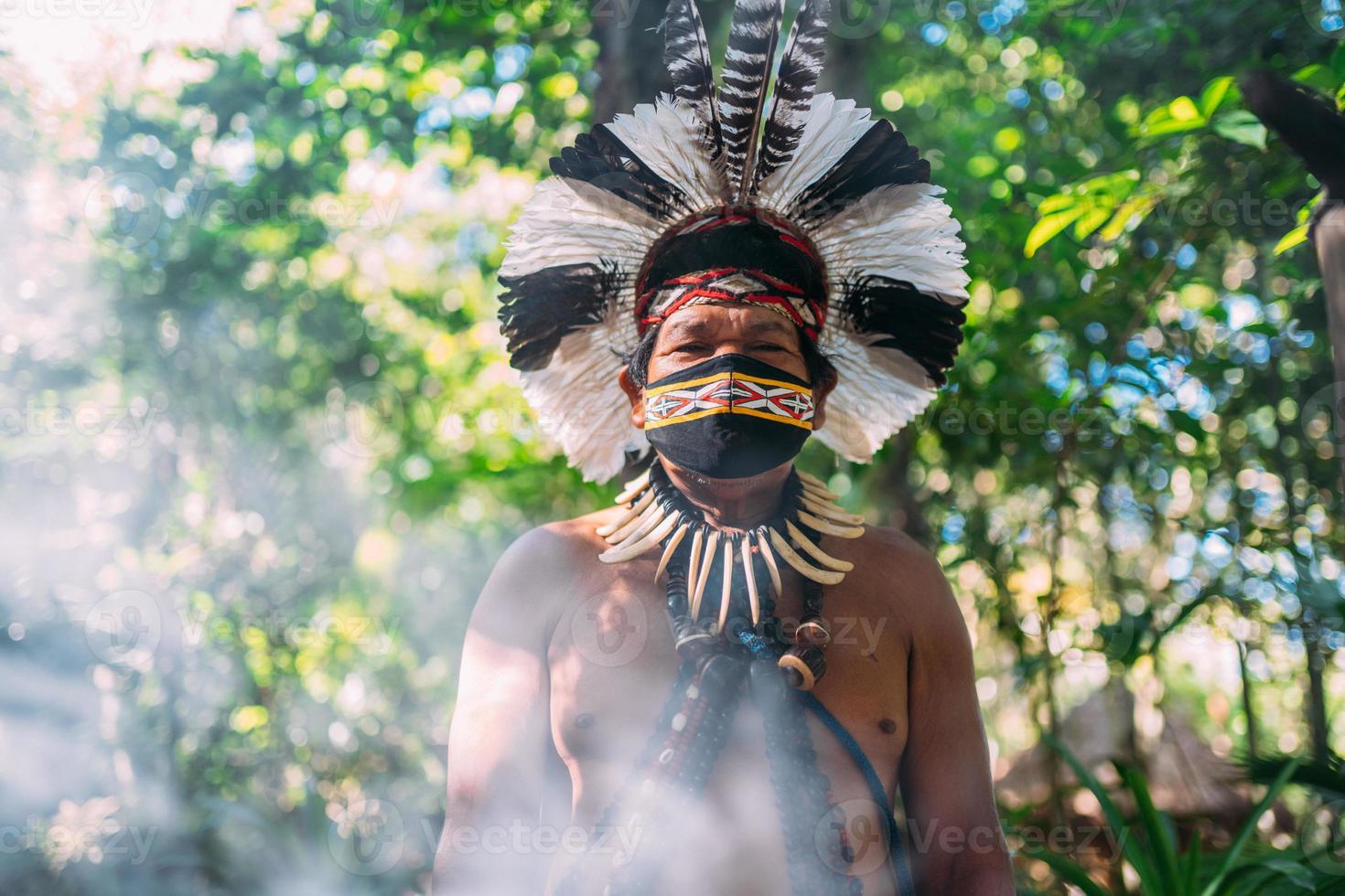 chamán de la tribu pataxo. anciano indio con tocado de plumas y máscara facial debido a la pandemia del covid-19. indio brasileño mirando a la cámara foto