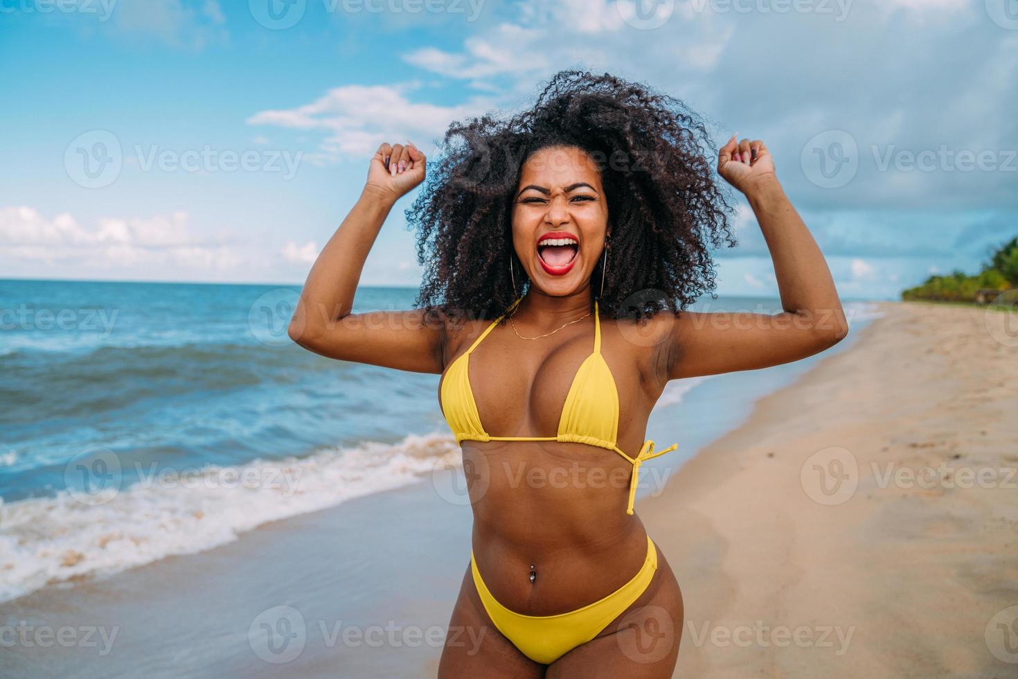 hermosa mujer hispana en bikini en la playa. mujer joven disfrutando de sus vacaciones de verano en un día soleado, sonriendo y celebrando mirando la cámara foto