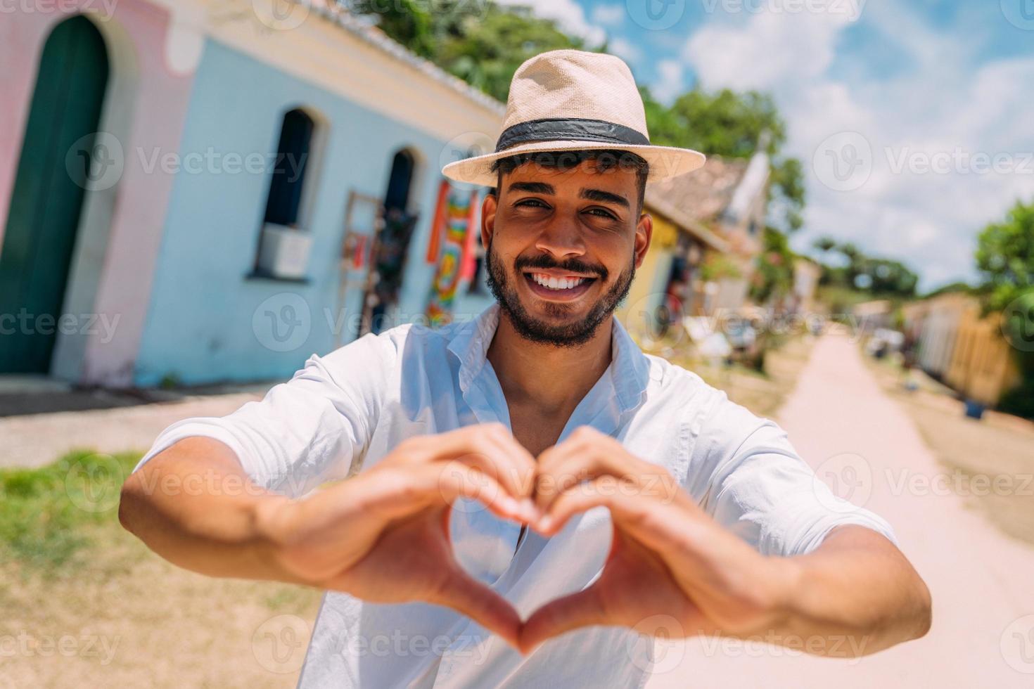 Tourist making selfie in the historic center of Porto Seguro. Latin American man in a hat, making a heart with his hands, smiling at the camera photo