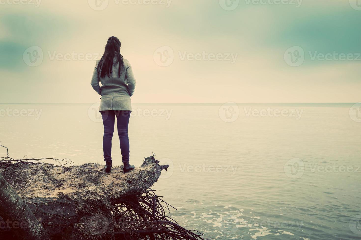 Woman standing on broken tree on wild beach looking at sea horizon photo