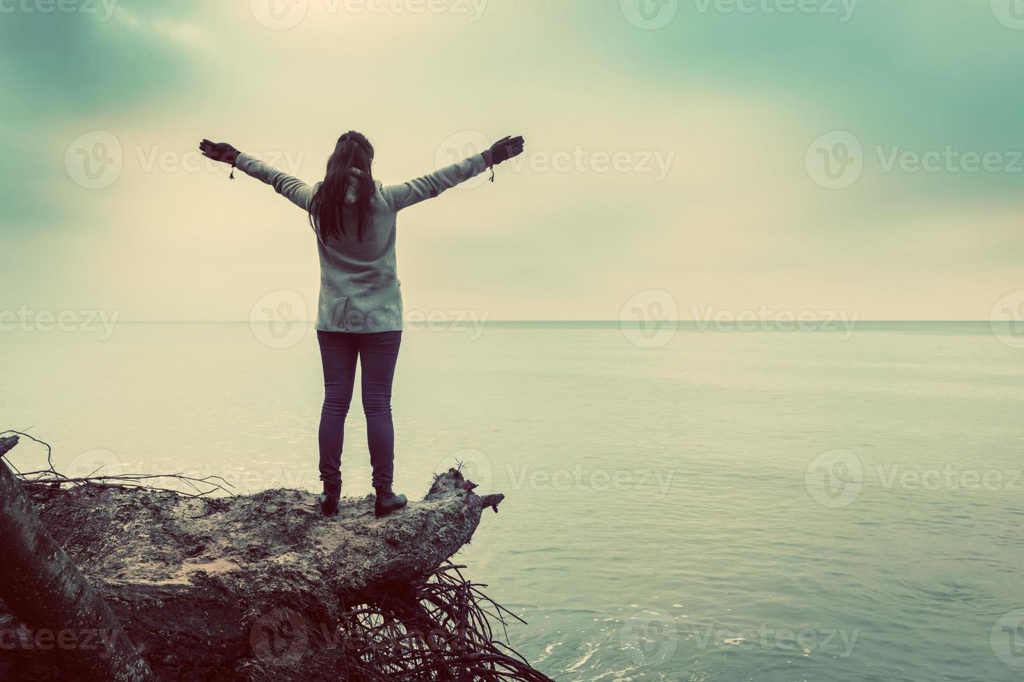 Woman standing on broken tree on wild beach with arms raised looking at sea photo