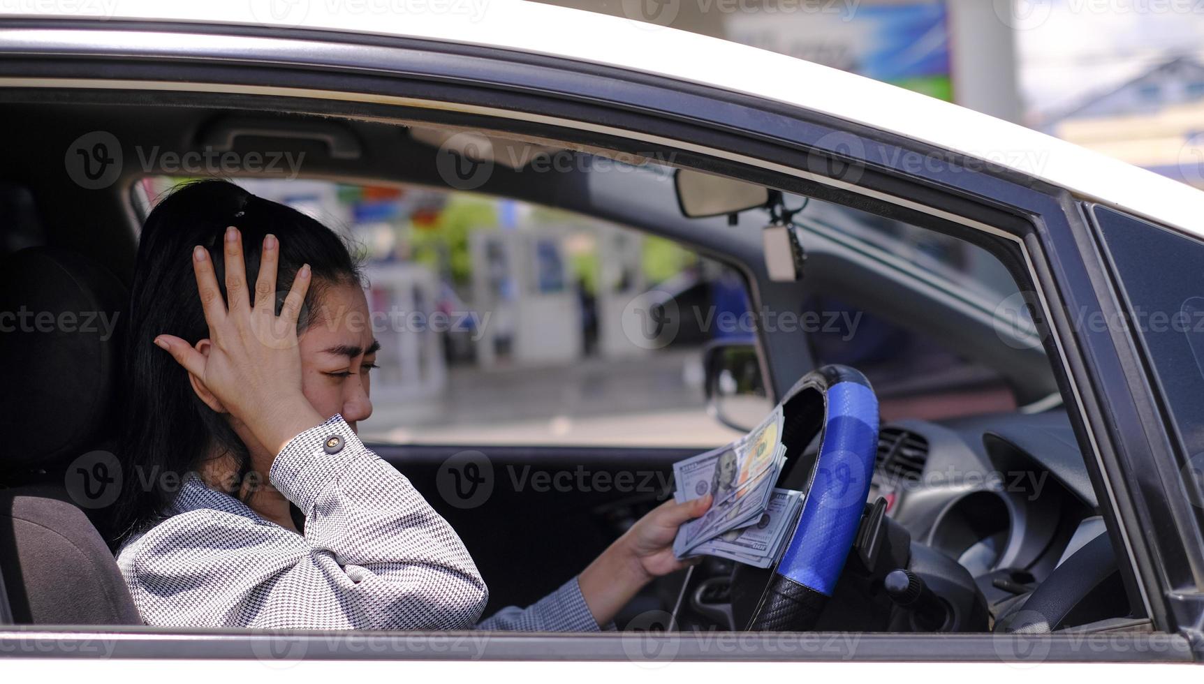 Desperate beautiful Asian woman holds cash dollar bills sitting inside her car at a gas station photo