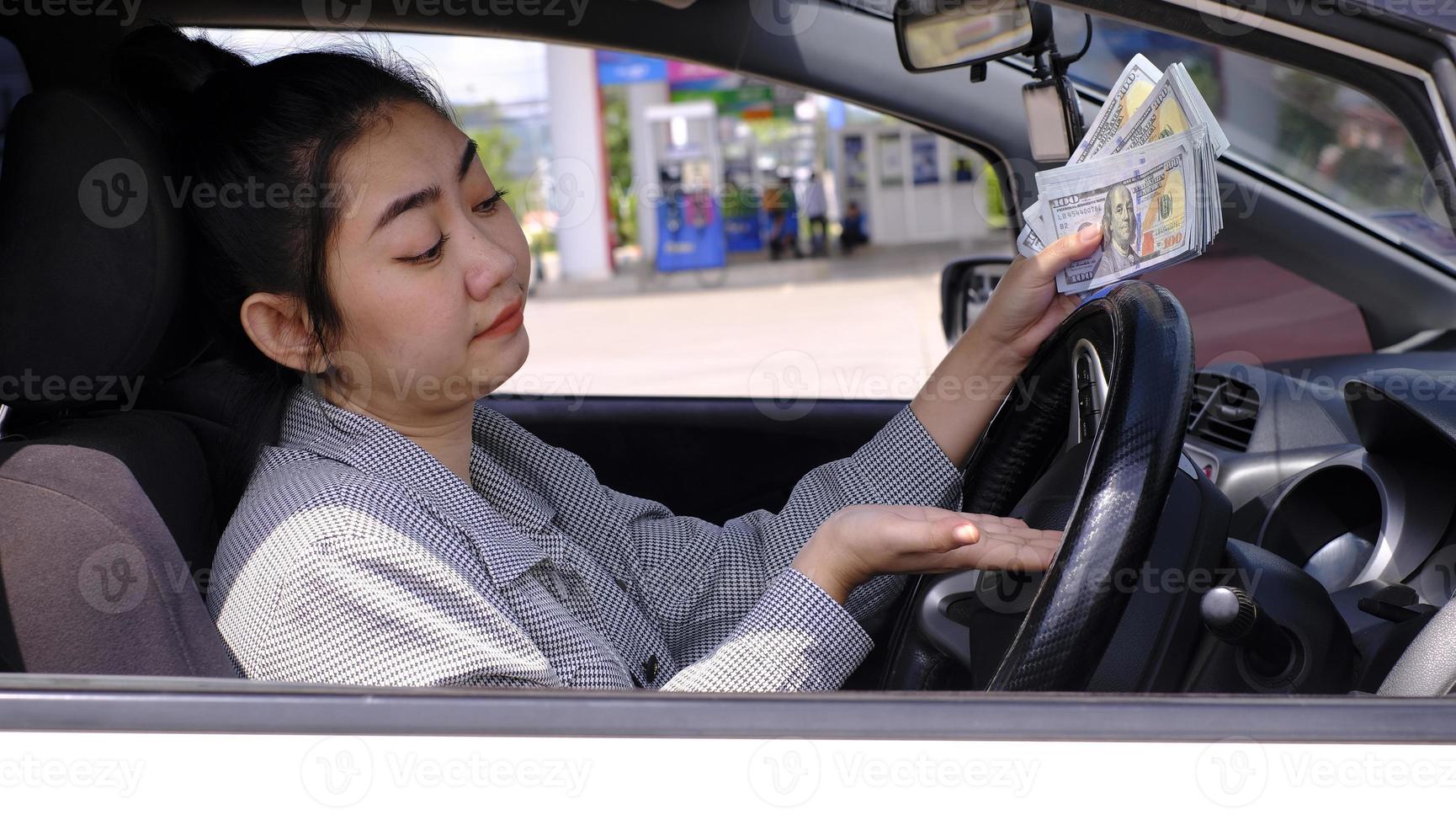 Desperate beautiful Asian woman holds cash dollar bills sitting inside her car at a gas station photo